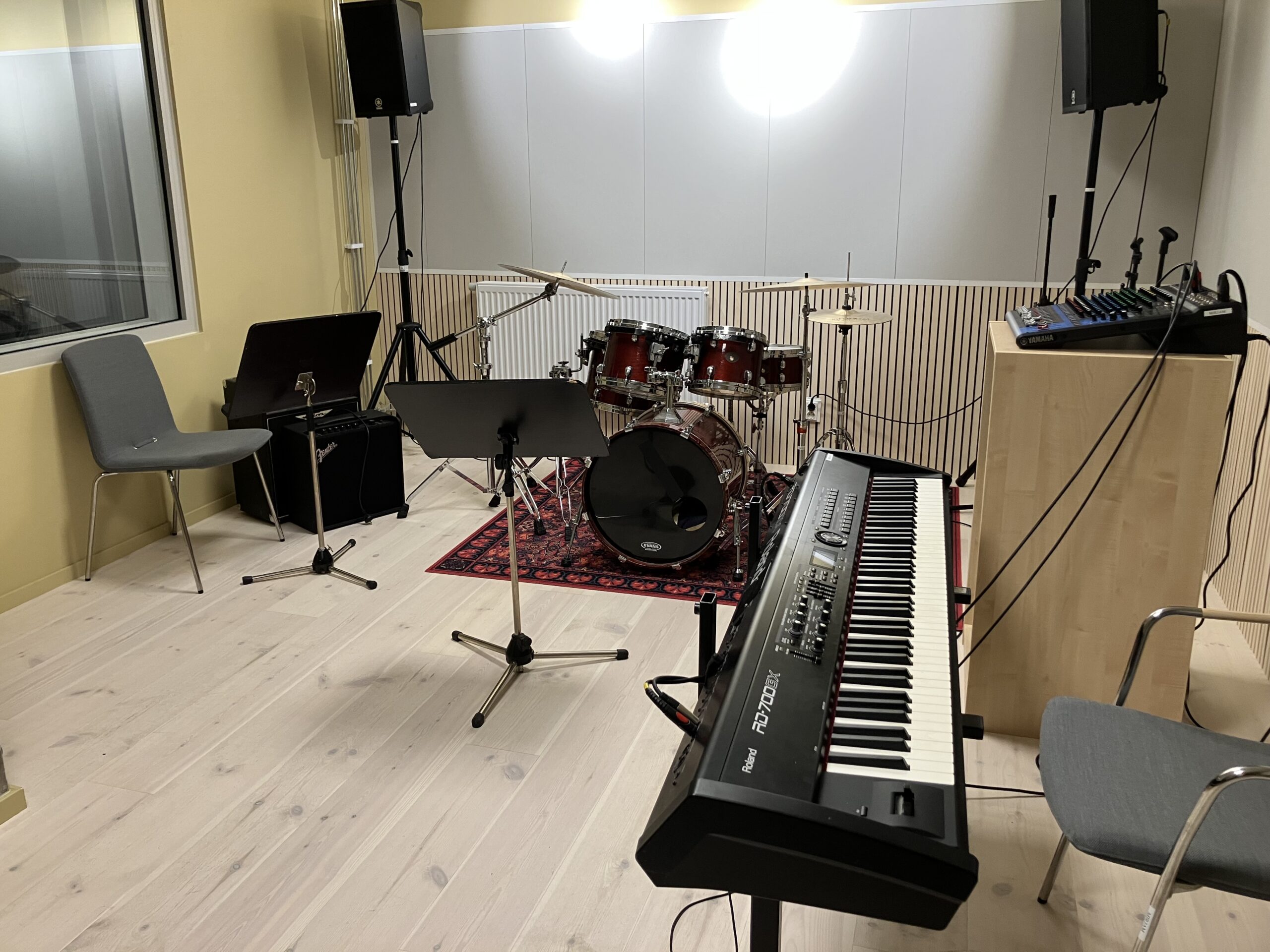 Ett musikrepetitionsrum med trumset, keyboard, notställ, mixerbord, förstärkare, högtalare och stolar placerade på ett ljust trägolv.