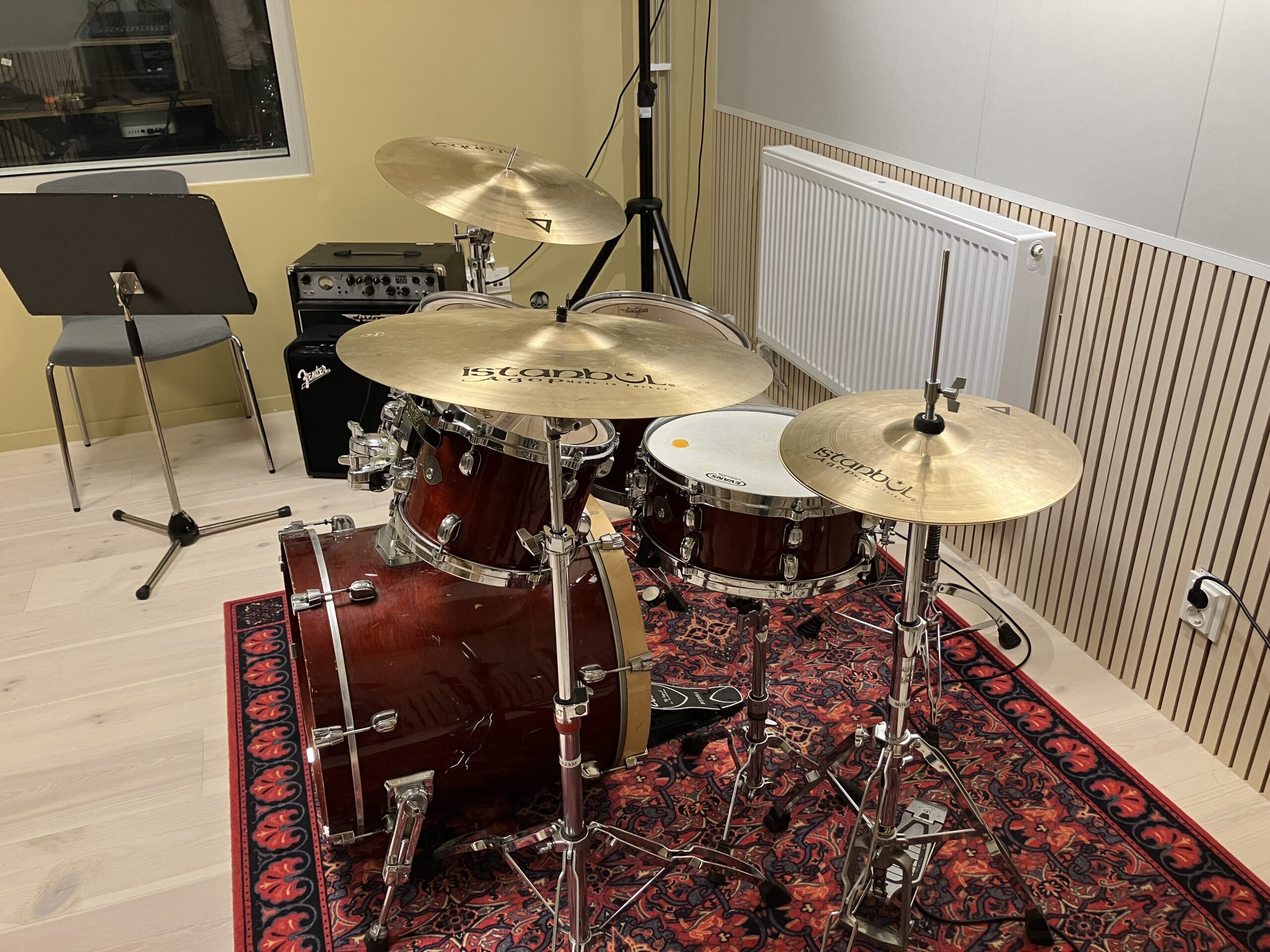 Ett trumset med cymbaler och bastrumma är placerat på en rödmönstrad matta i ett musikövningsrum med notställ, förstärkare och element i närheten.