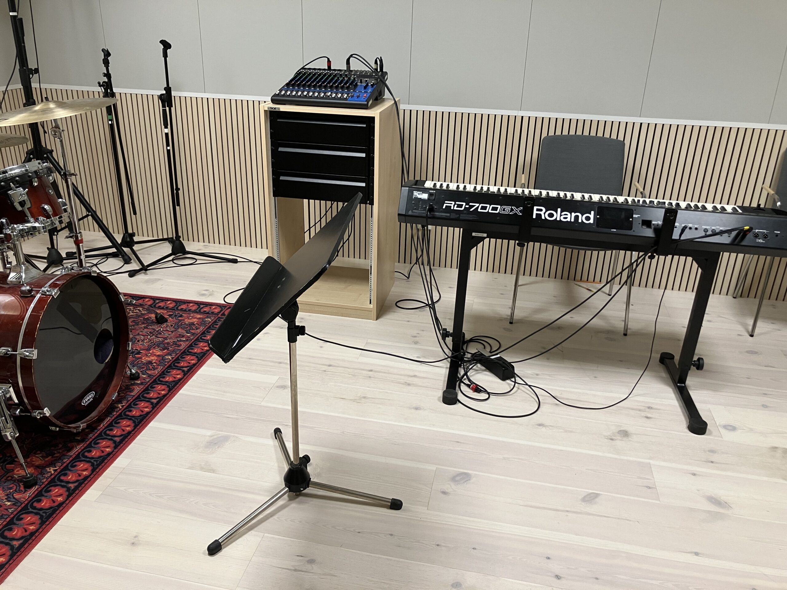 Ett musikövningsrum med ett trumset, notställ, Roland RD-700GX keyboard, ljudmixer på en hylla och en stol på ett ljust trägolv.
