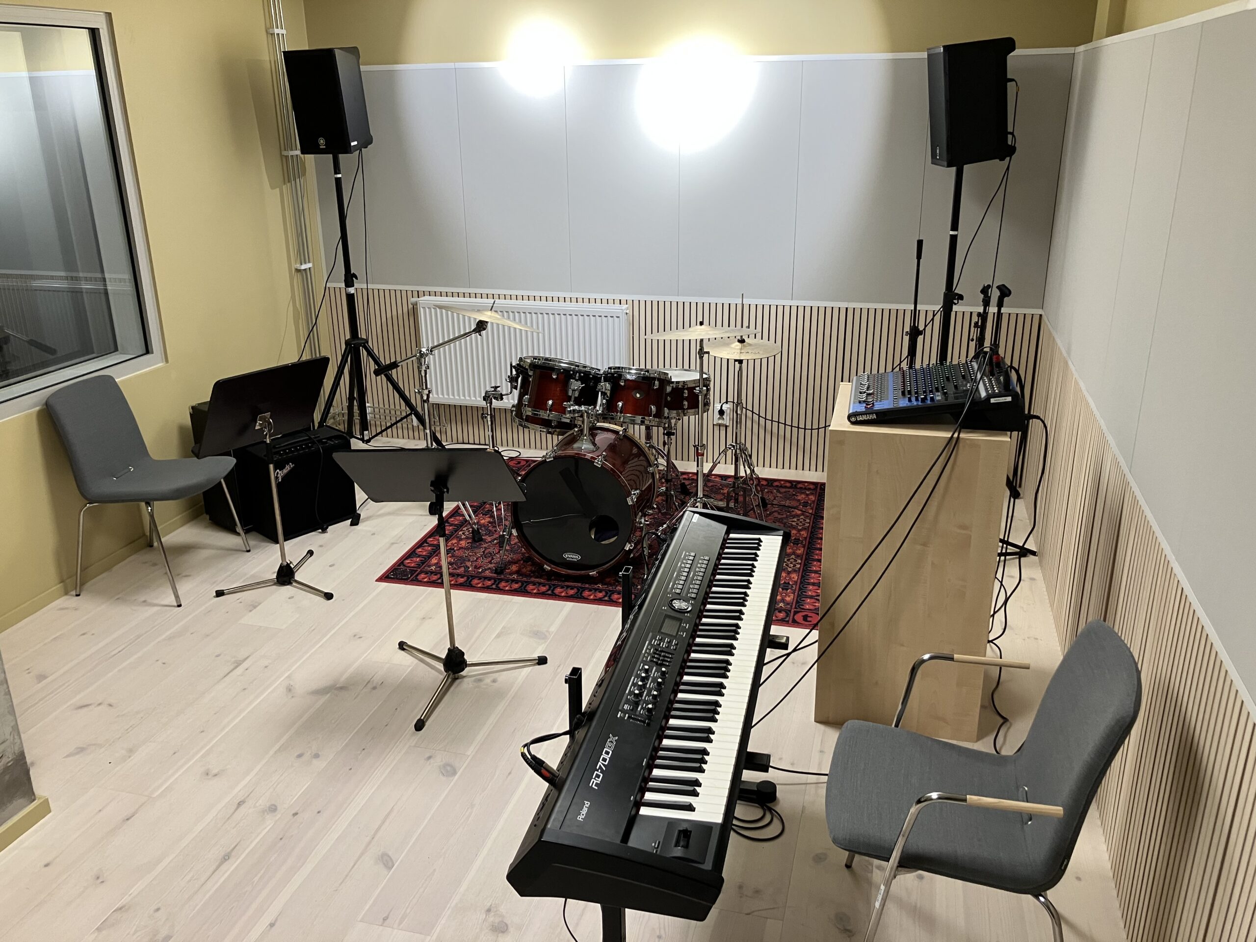 Ett musikövningsrum med ett trumset, keyboard, två stolar, notställ, högtalare och ljudutrustning på ett ljust trägolv.