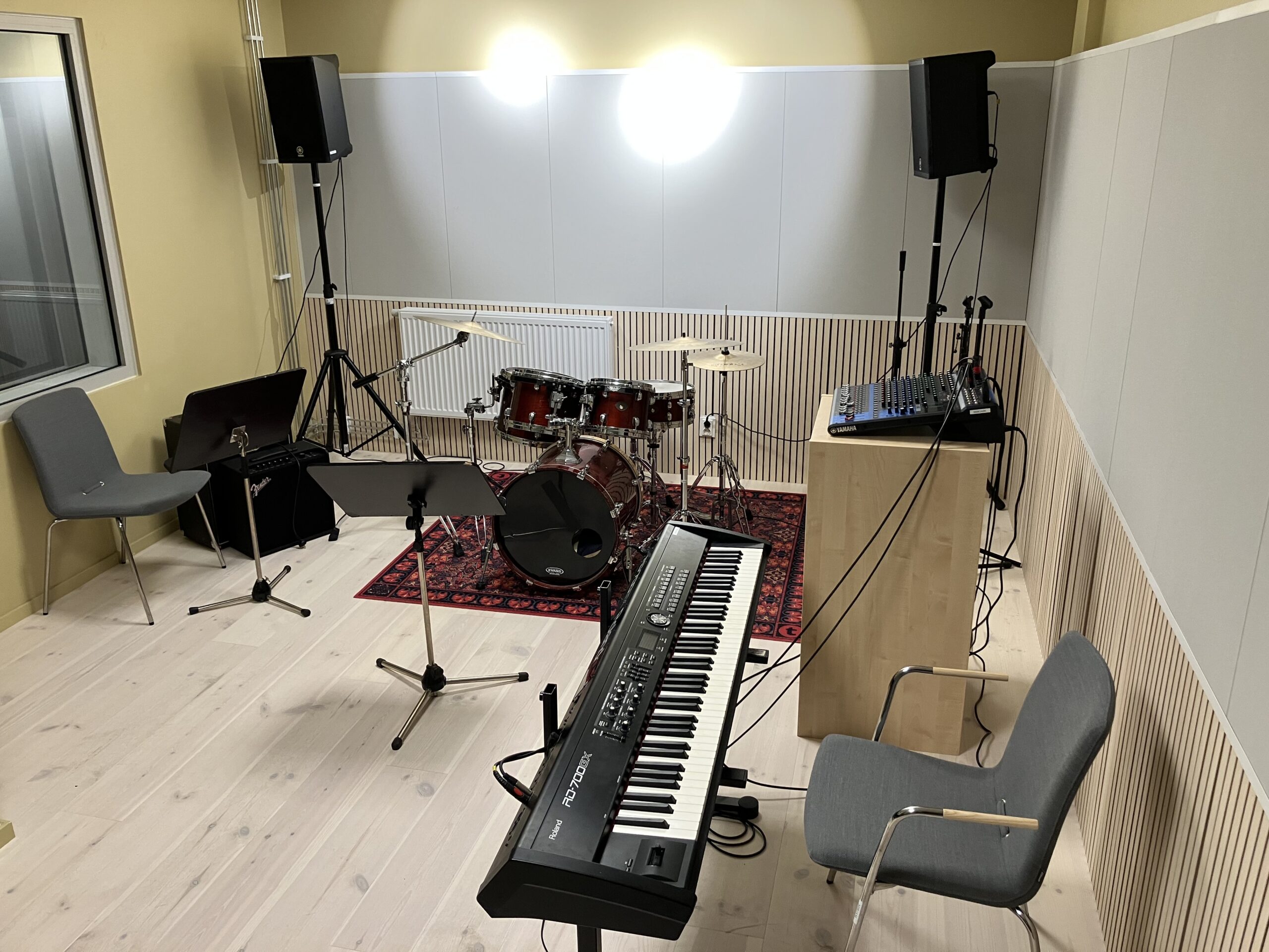 En liten musikstudio med trumset, keyboard, notställ, mixerbord på ett bord, stolar och två högtalare på stativ.