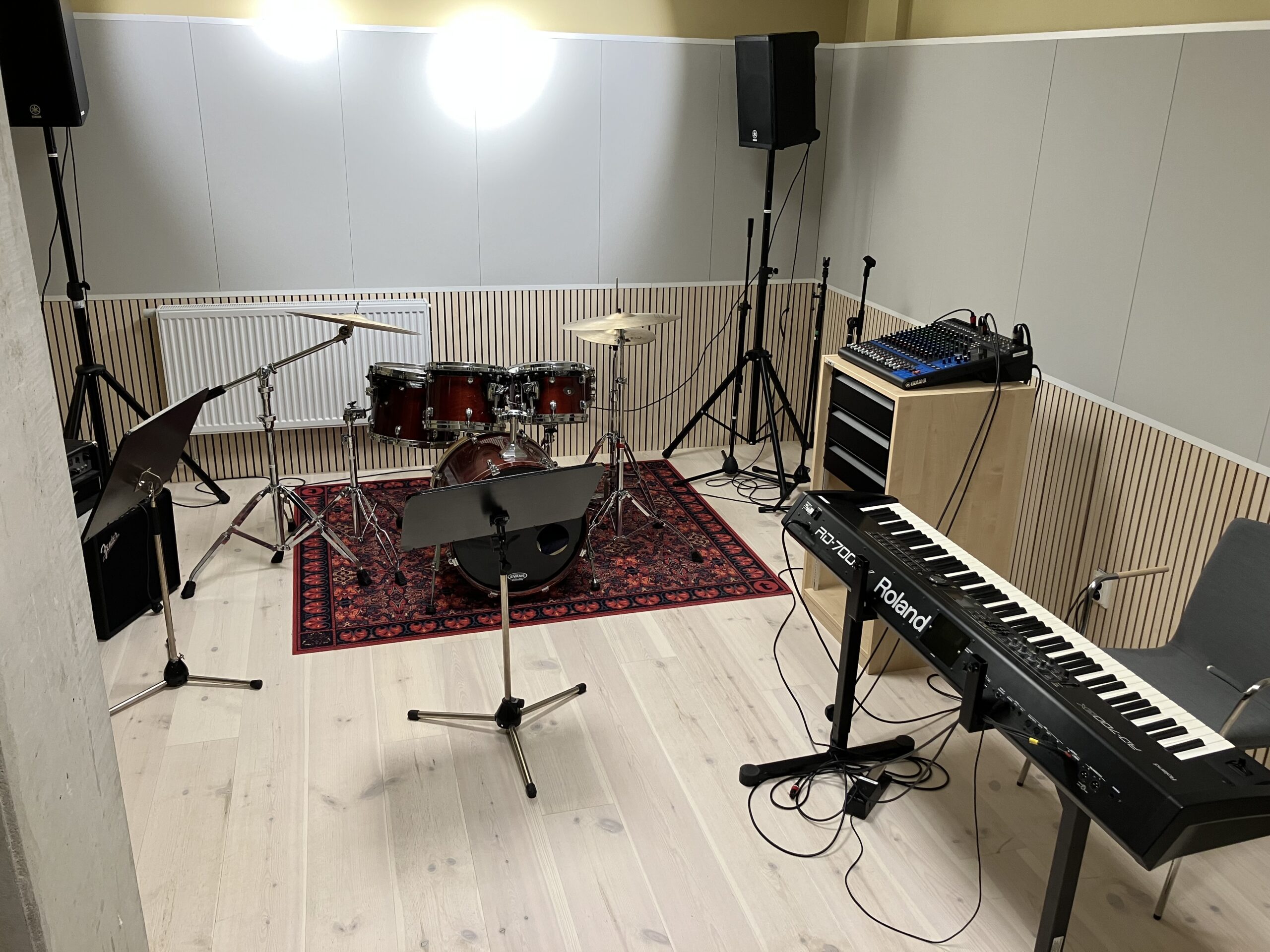 Ett musikrepetitionsrum med trumset, keyboards, notställ, mikrofoner, högtalare, mixerbord och en rödmönstrad matta.