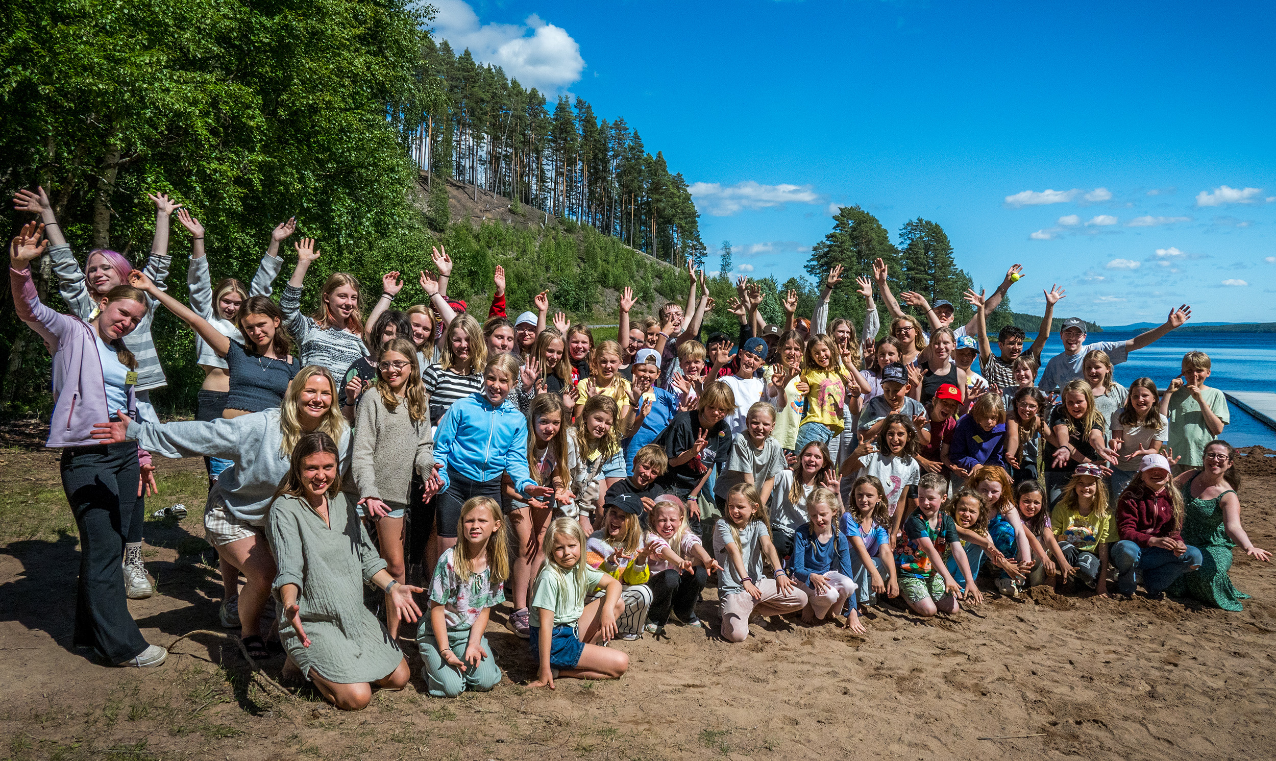 En stor grupp människor, inklusive barn, poserar och vinkar glatt på en sandstrand med träd och en sjö i bakgrunden under en klarblå himmel.