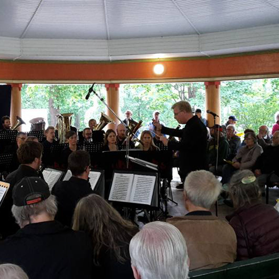 En dirigent leder en bandensemble i ett utomhuslusthus medan en publik lyssnar uppmärksamt, några sittande och andra stående.