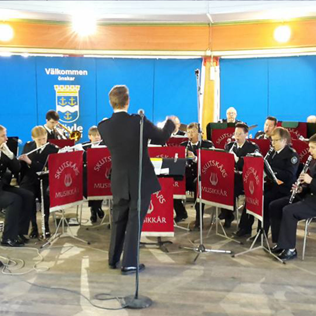 En dirigent leder ett gäng musiker som spelar blås- och träblåsinstrument på en scen med blå bakgrund och vapen.