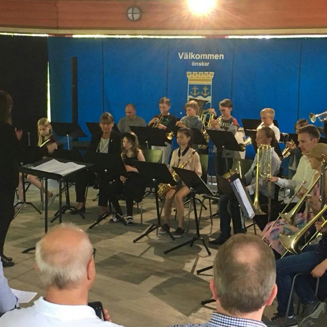 En grupp musiker, inklusive barn och vuxna, uppträder inför publik. En dirigent står till vänster medan en blå banderoll med text på svenska hänger bakom artisterna.