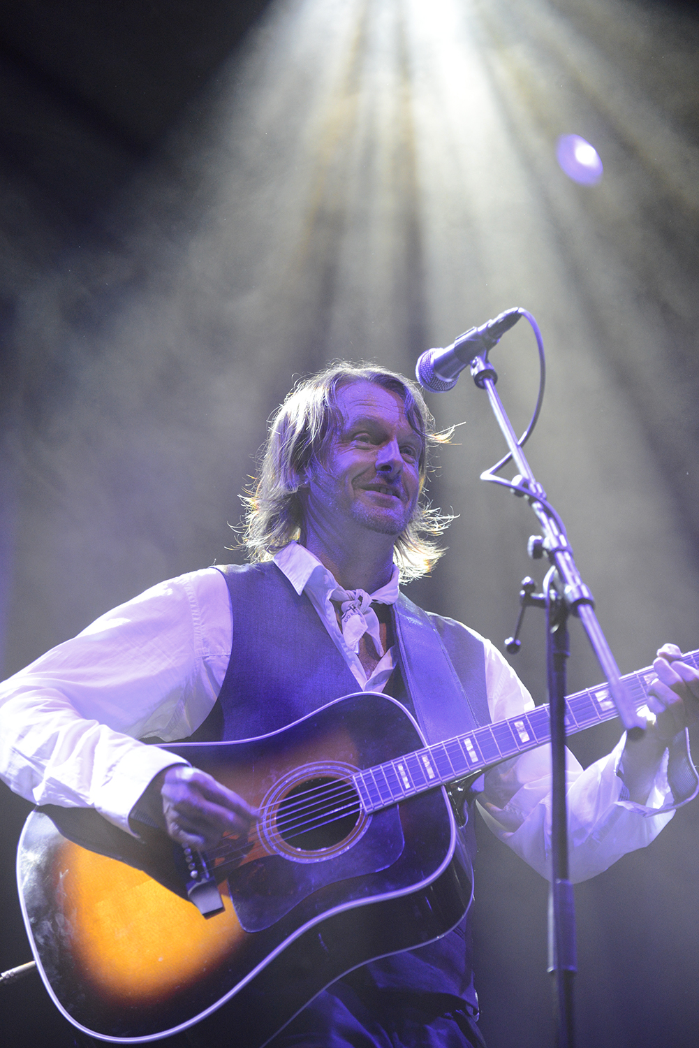 En person står på scenen och spelar en akustisk gitarr under en strålkastare, iklädd en vit skjorta, slips och väst.