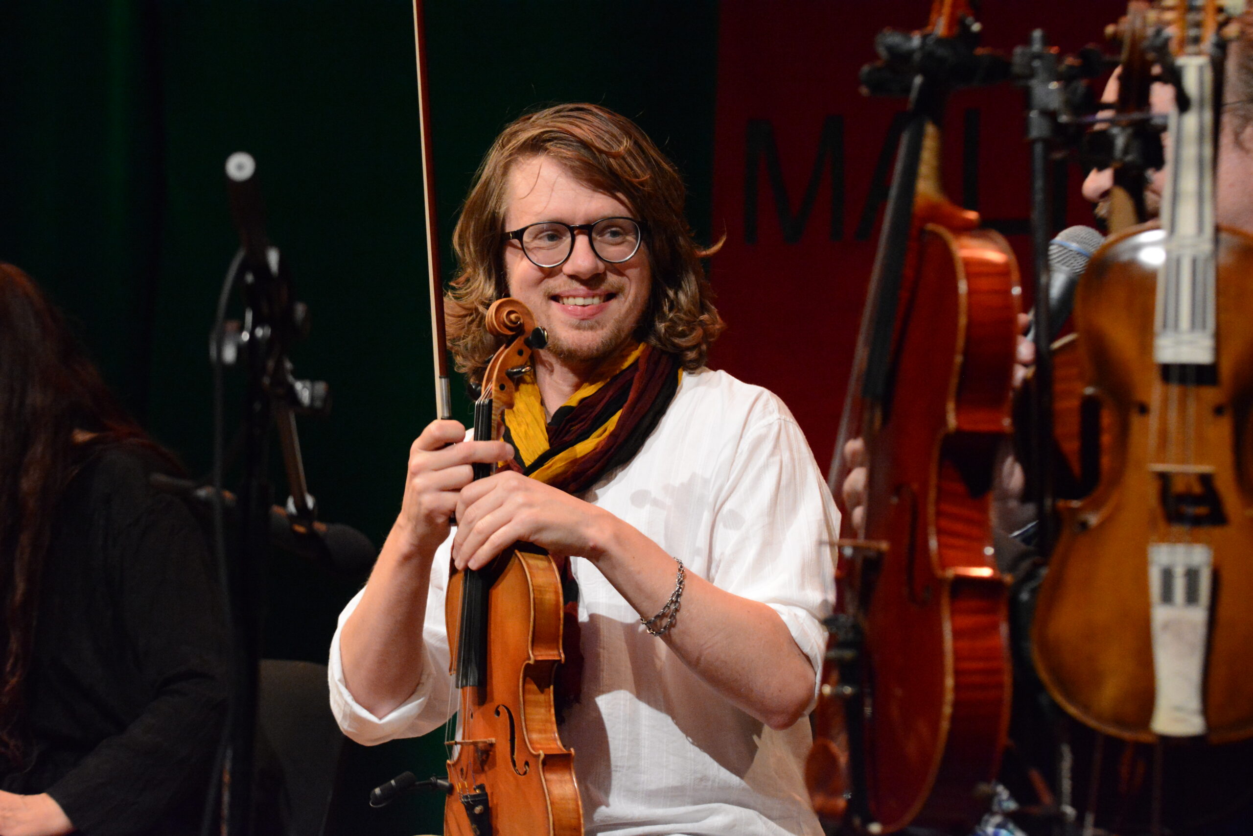 En person med glasögon håller en fiol och stråke, sittande bland andra instrument på en scen.