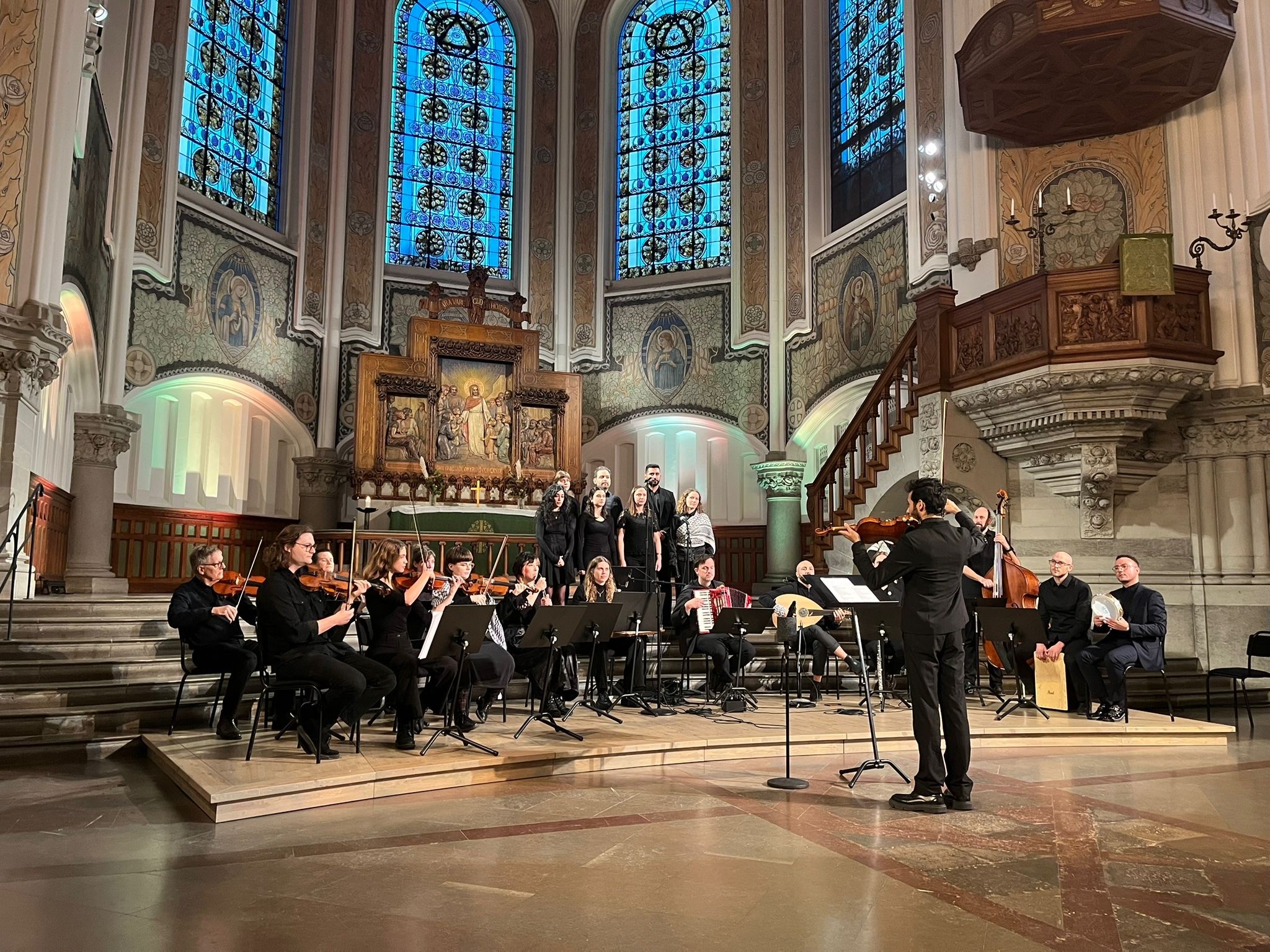 En orkester och kör uppträder i en historisk kyrka med glasmålningar och utsmyckade dekorationer.