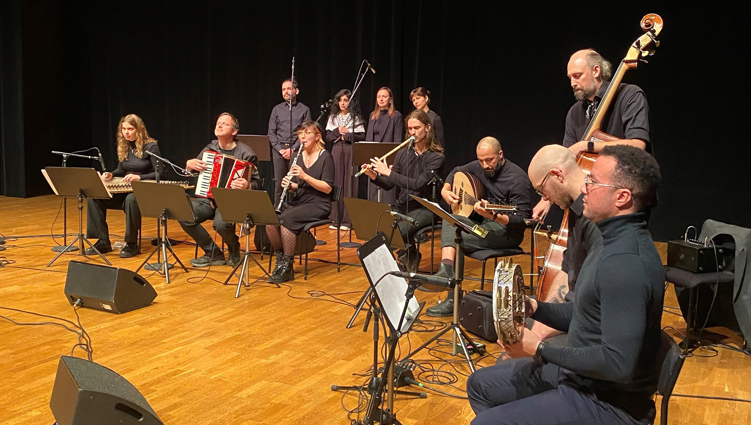 En ensemble av musiker som sitter på en scen och spelar olika instrument inklusive dragspel, flöjt, gitarr och kontrabas. Framför dem står mikrofoner och notställ.