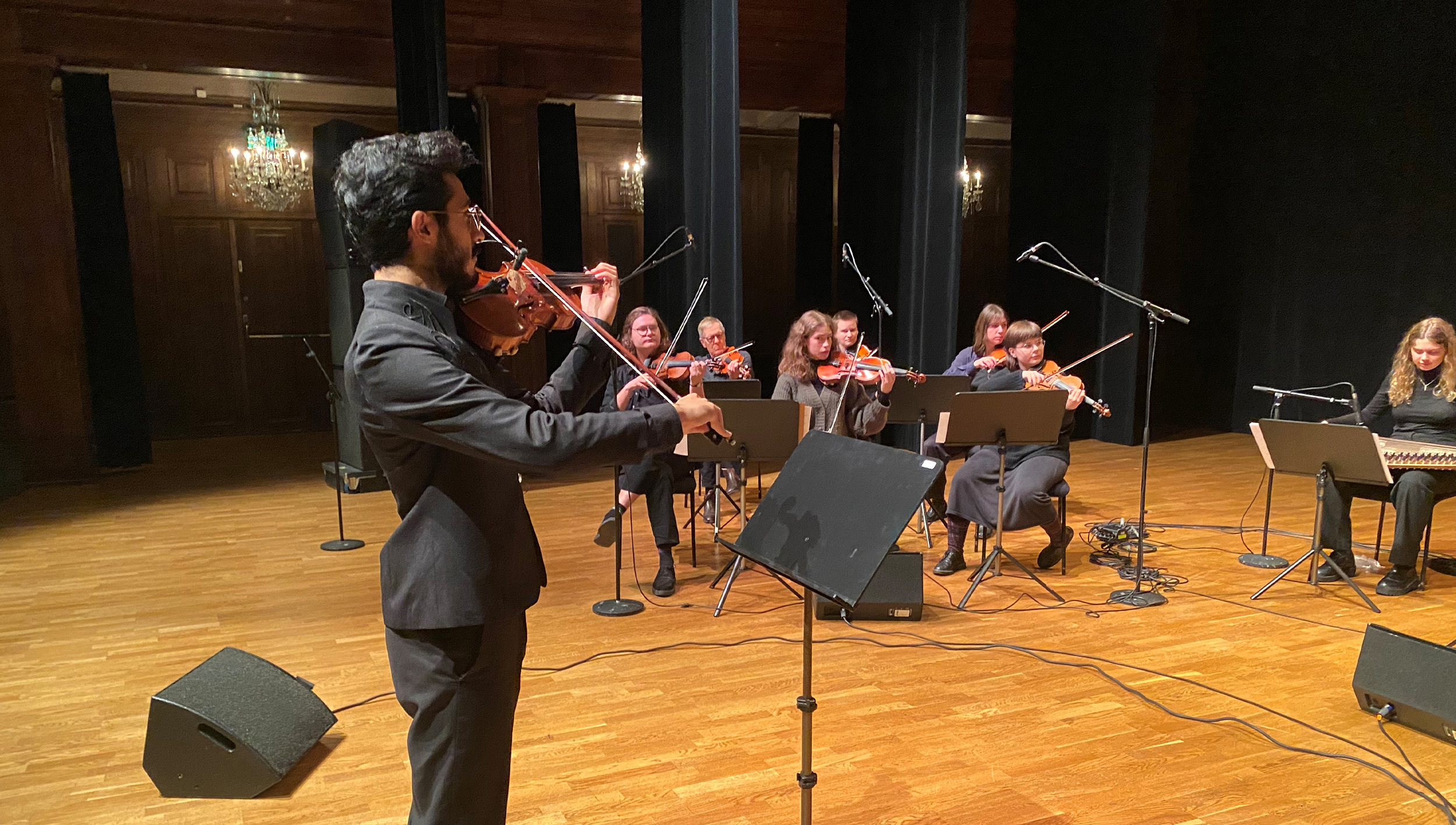 En dirigent spelar fiol framför en orkester som sitter på en träscen.