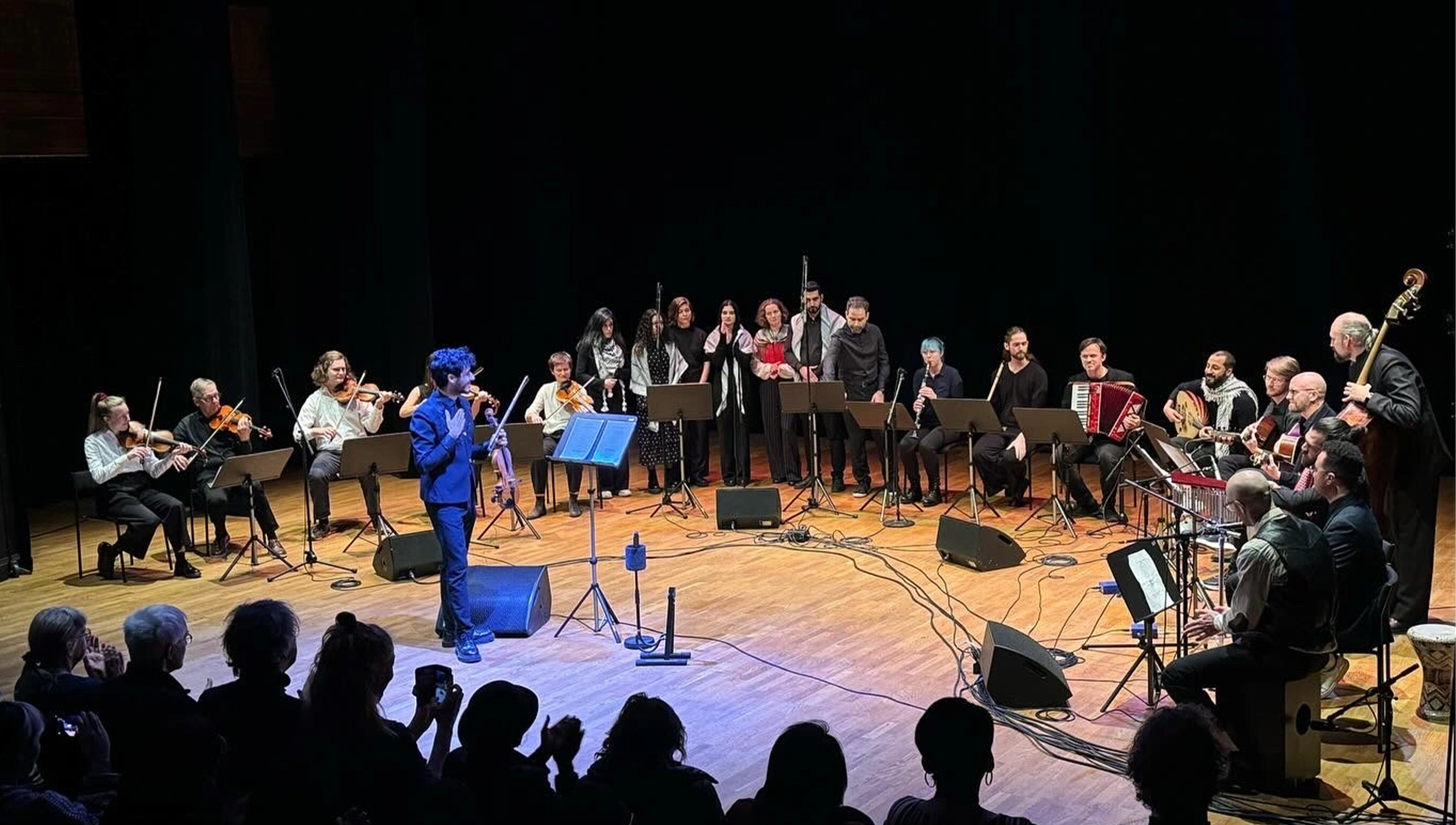 En dirigent leder en orkester och kör på scenen under en föreställning. Musiker spelar olika instrument och publiken syns i förgrunden.