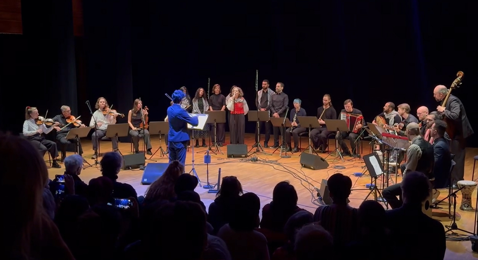 En dirigent leder en orkester och kör på scenen under en föreställning, med sittande publik i förgrunden.