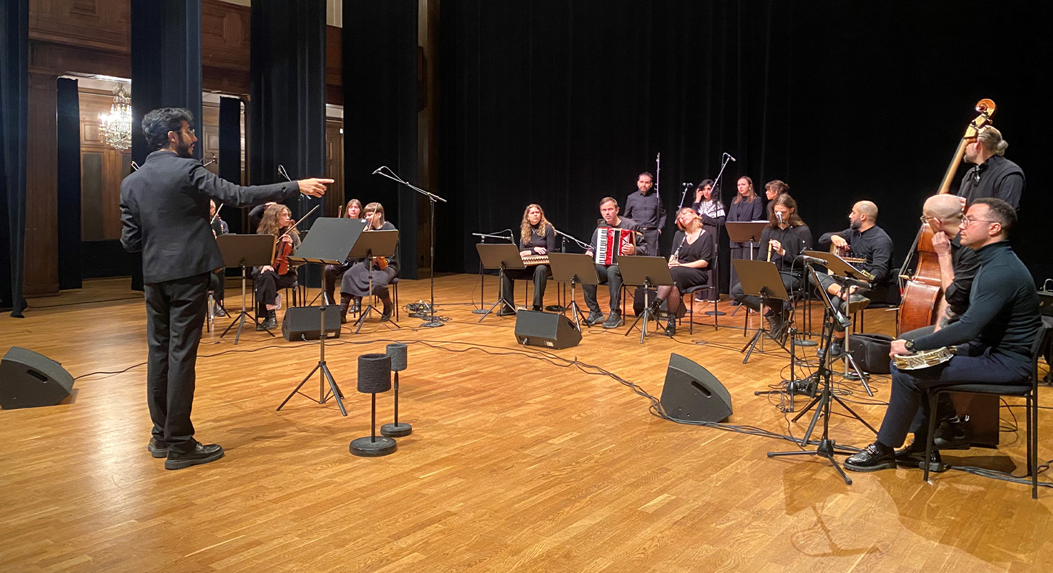 Dirigent leder en ensemble musiker på scenen, med olika stråk-, blås- och dragspelare, med mikrofoner inställda för ljud.