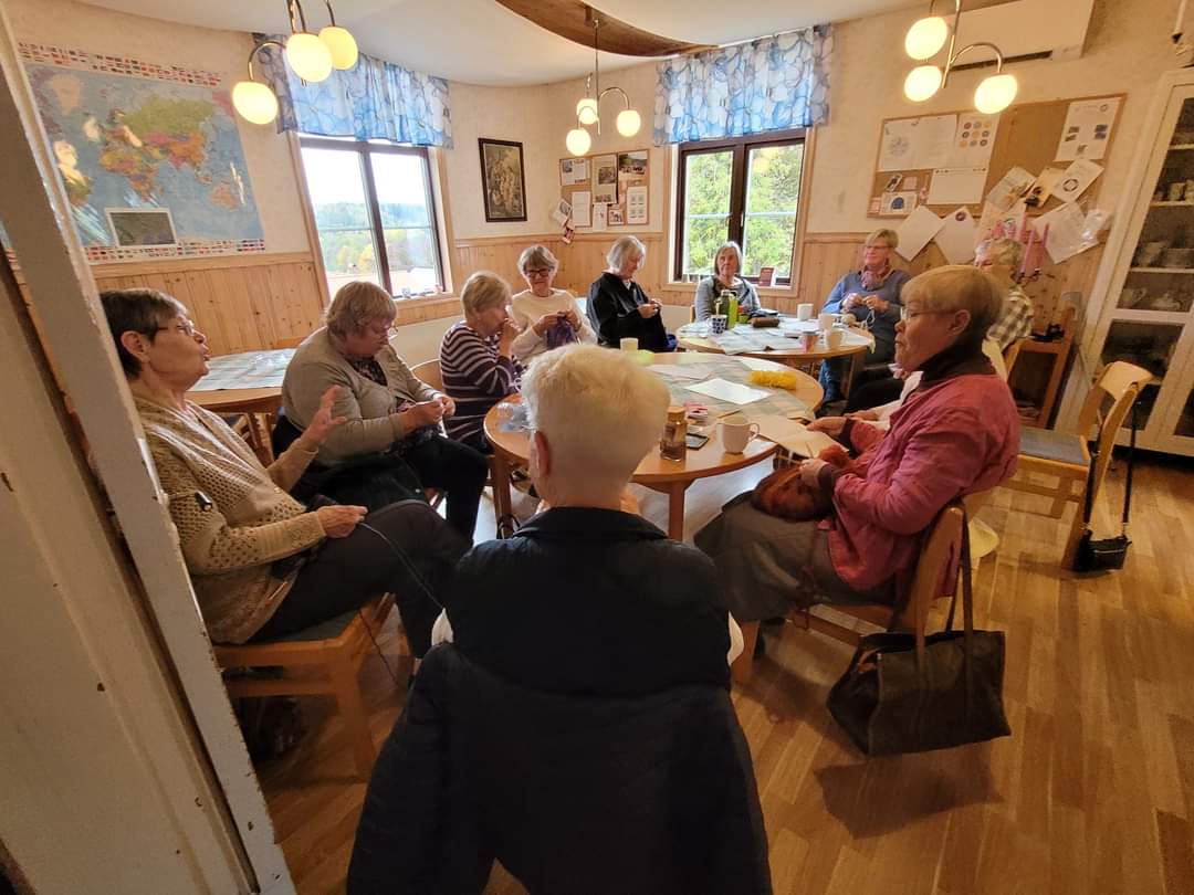 En grupp äldre kvinnor sitter runt ett bord och stickar i ett mysigt rum med kartor och affischer på väggarna.