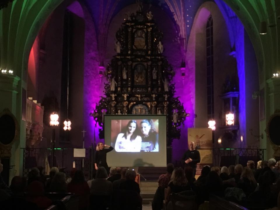 En presentation äger rum i en svagt upplyst kyrka med ett utsmyckat altare. En stor skärm visar en bild för en publik som sitter i bänkar.