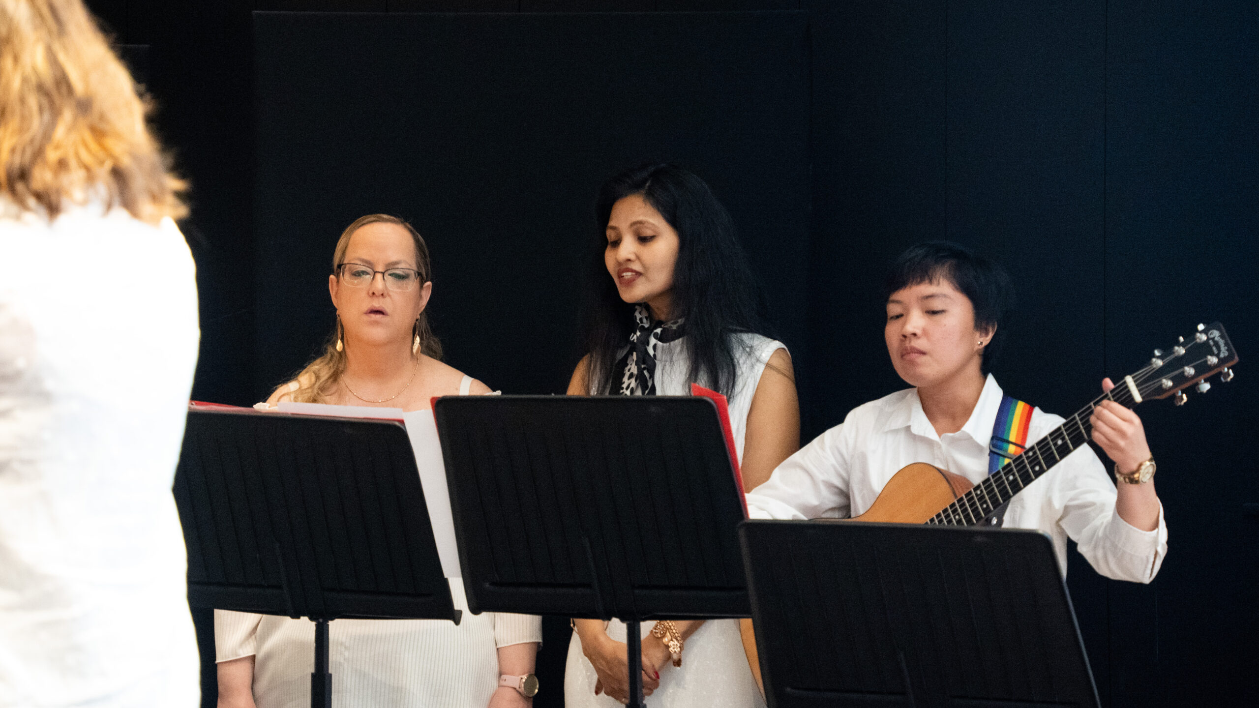 Tre personer står bakom notställ; två sjunger medan en spelar gitarr. Alla är klädda i ljusa kläder och uppträder inomhus mot en mörk bakgrund.