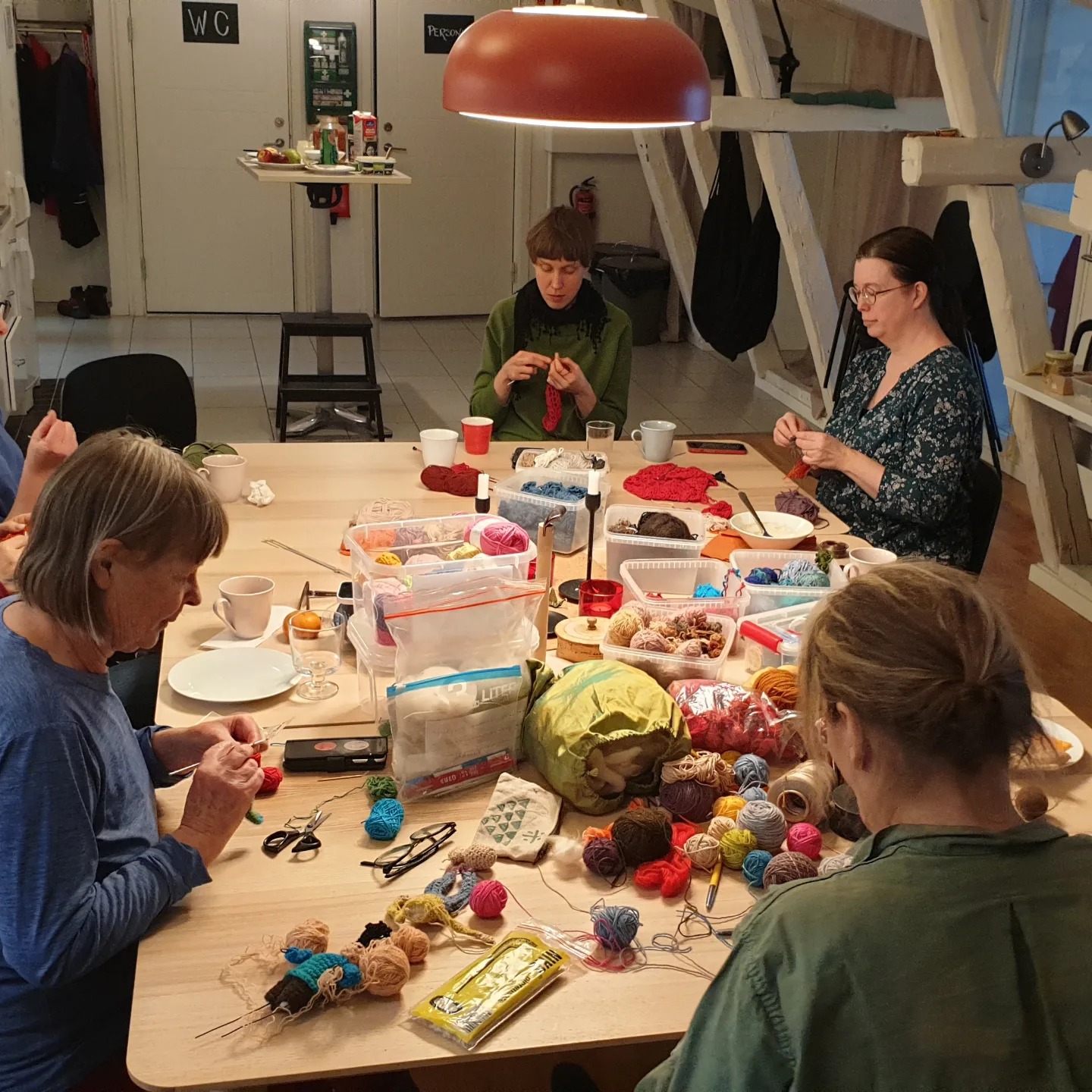Fem personer sitter runt ett bord täckt med garn, pysselmaterial och snacks och ägnar sig åt stickning och andra pysselaktiviteter i ett väl upplyst rum.