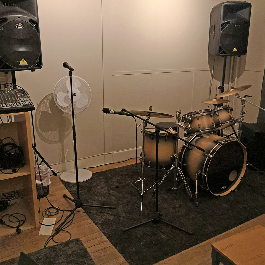 Ett trumset, två mikrofoner på stativ, ett mixerbord, en fläkt och två stora högtalare i ett litet musikrepetitionsrum.