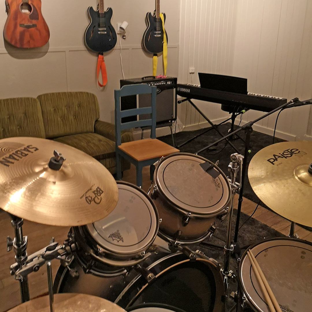 Ett trumset, en elektrisk keyboard, förstärkare och flera gitarrer på väggen i ett mysigt musikövningsrum med en grön soffa och en trästol.