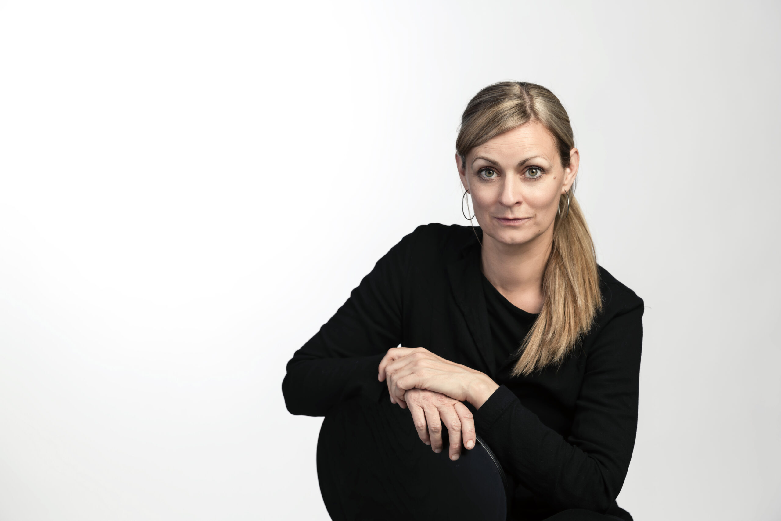 Kvinna med blont hår i en låg hästsvans, klädd i en svart långärmad topp, sitter med armarna vilande på knäna mot en vit bakgrund.