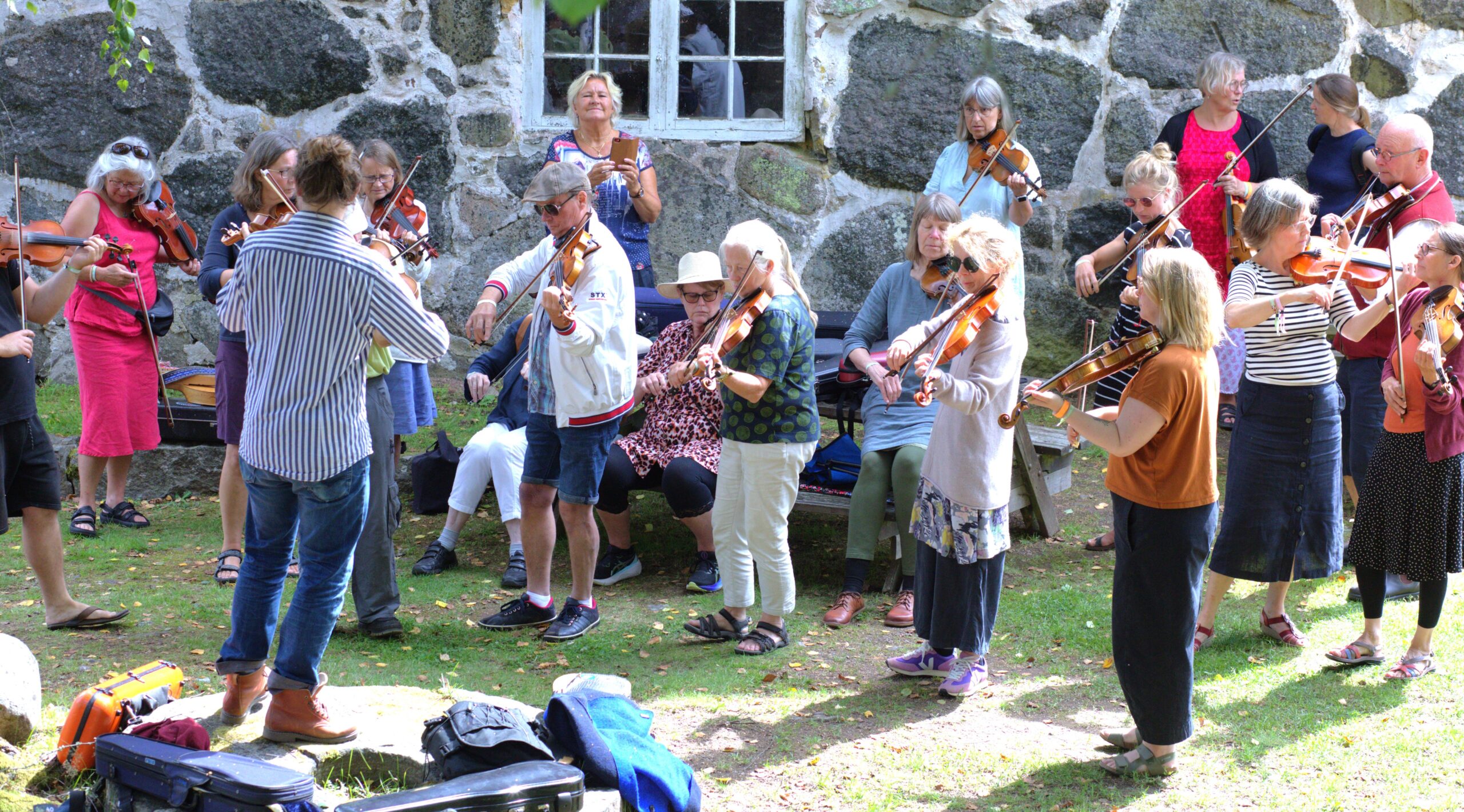 En grupp människor, de flesta stående, spelar violin utomhus framför en stenbyggnad, med instrumentfodral på marken i närheten.