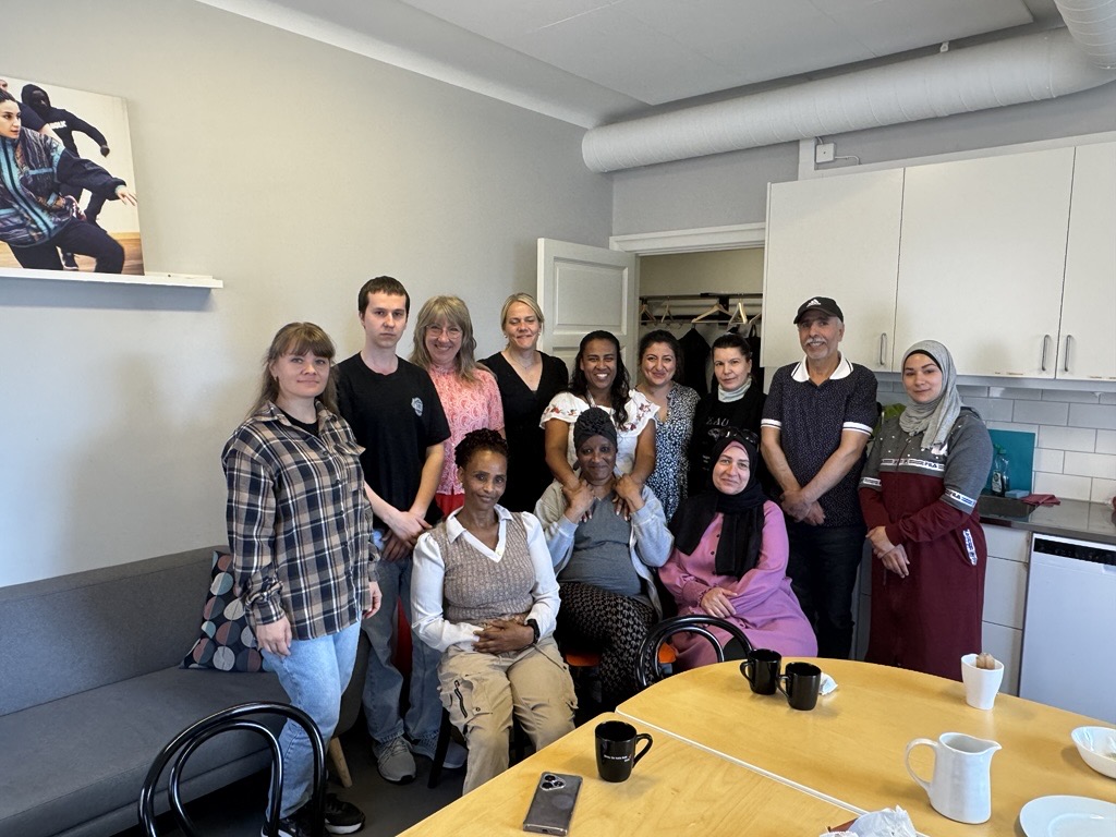 En grupp på tretton personer poserar och ler tillsammans i ett kök med muggar och tallrikar på bordet.