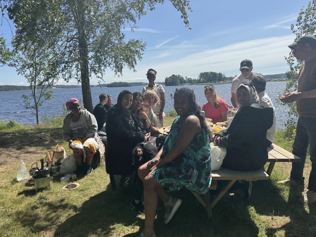 En grupp människor njuter av en picknick vid en sjö en solig dag, sittande och stående runt ett träbord.