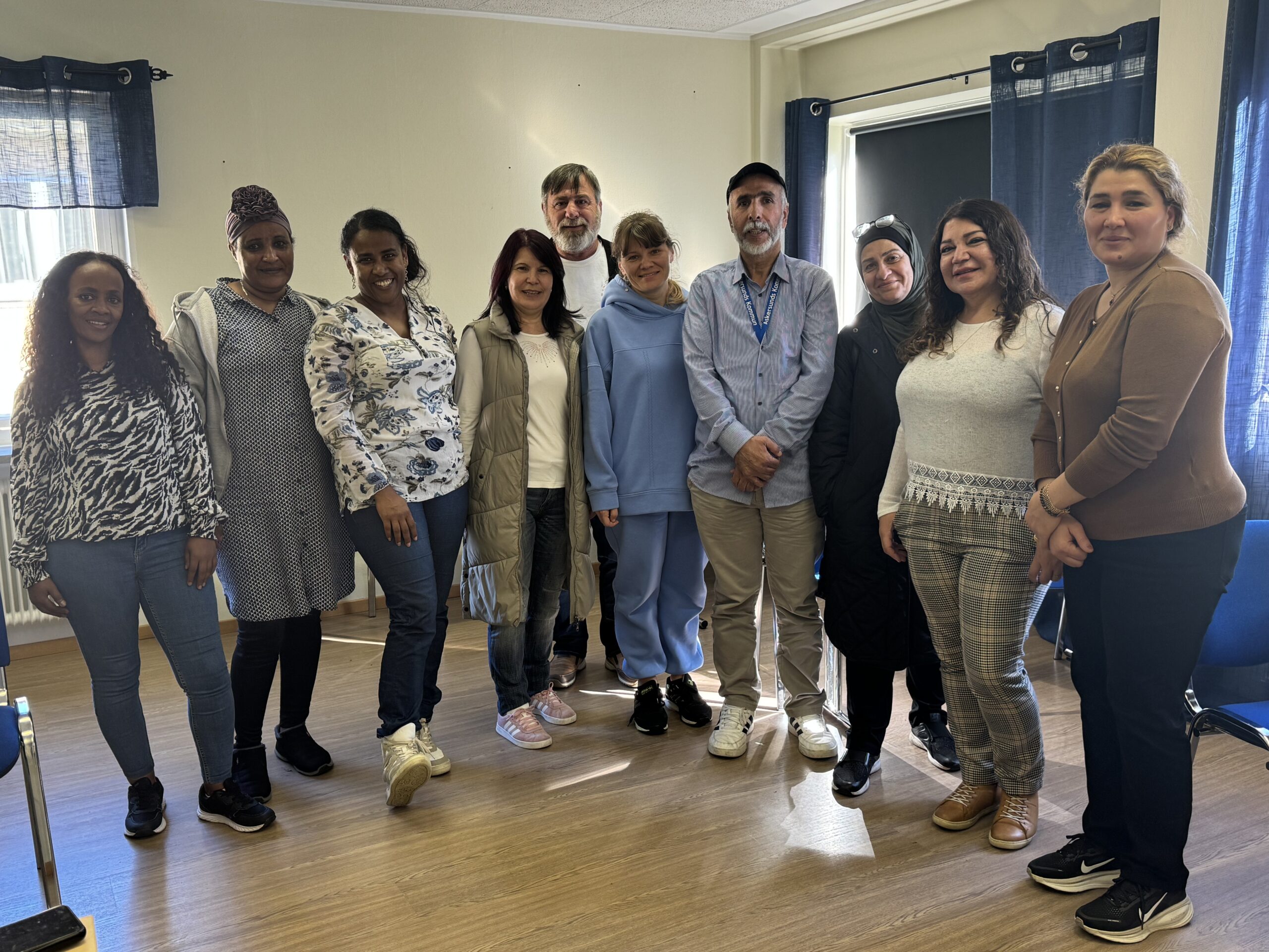 Tio personer står och ler tillsammans i ett rum med trägolv och blå gardiner.