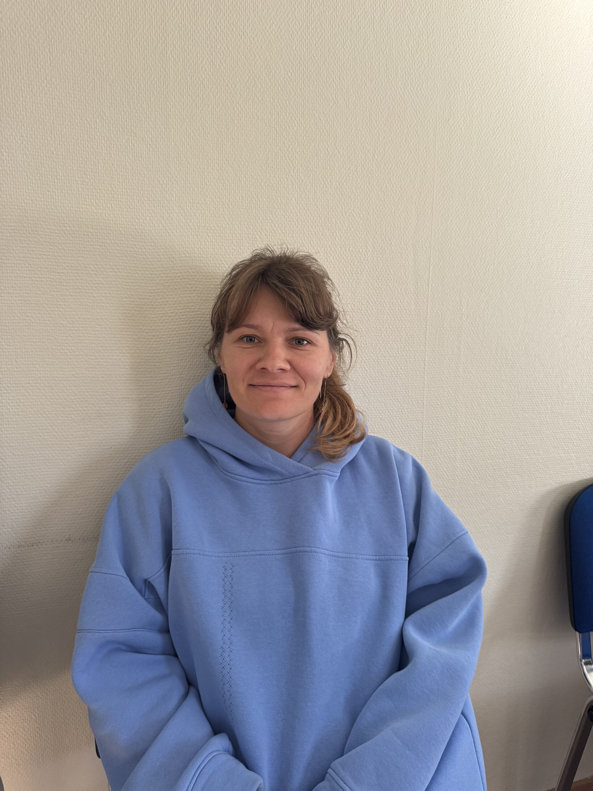 Kvinna med brunt hår i blå huvtröja sitter och ler mot en ljus vägg, bredvid en blå stol.
