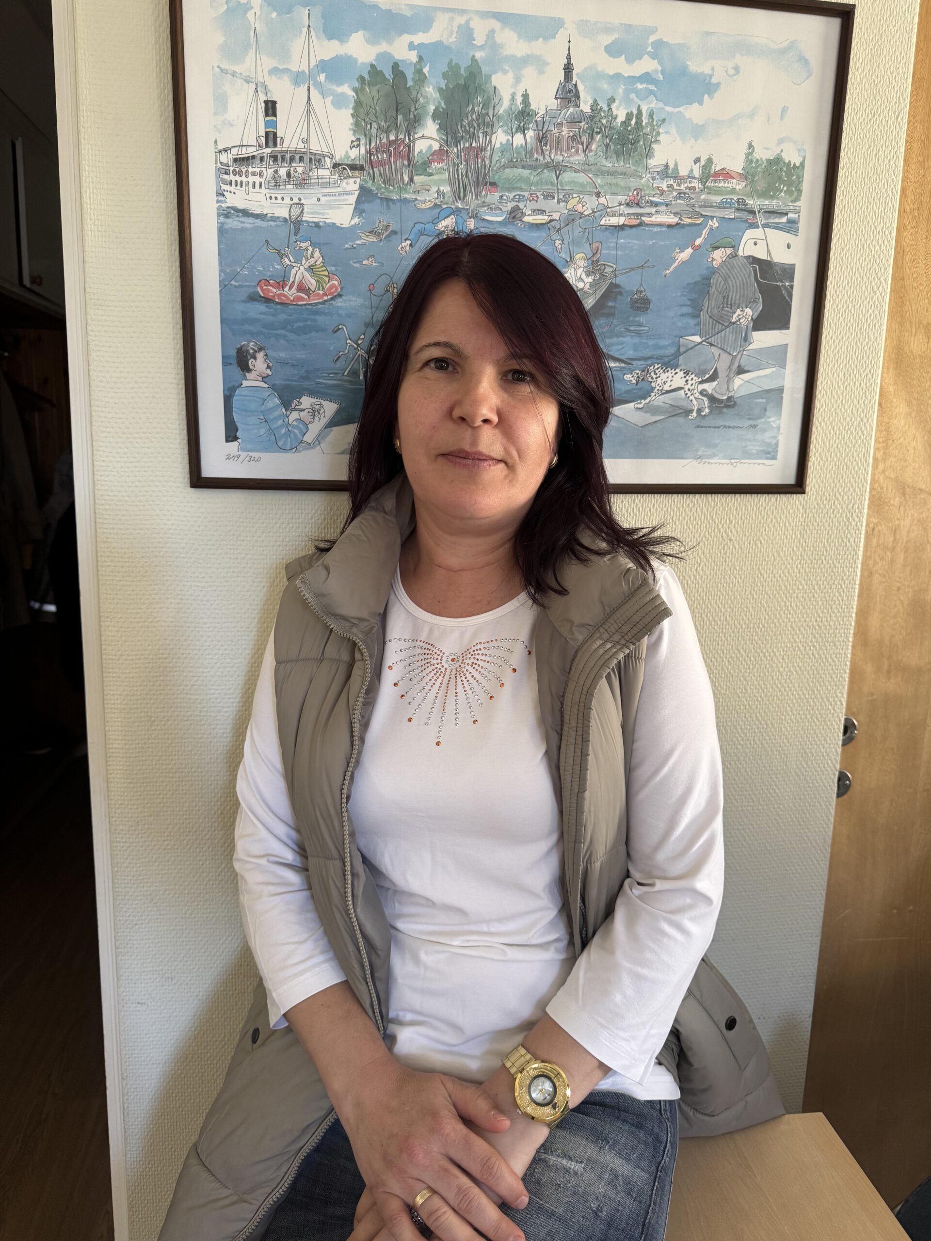 En kvinna med mörkt hår sitter inomhus framför en färgglad målning av båtar och människor vid vattnet.