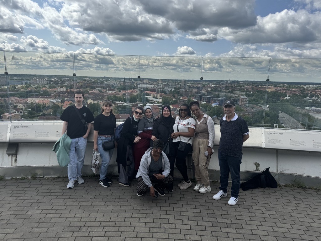 En grupp människor poserar tillsammans på ett tak med en stadsbild och en molnig himmel i bakgrunden.