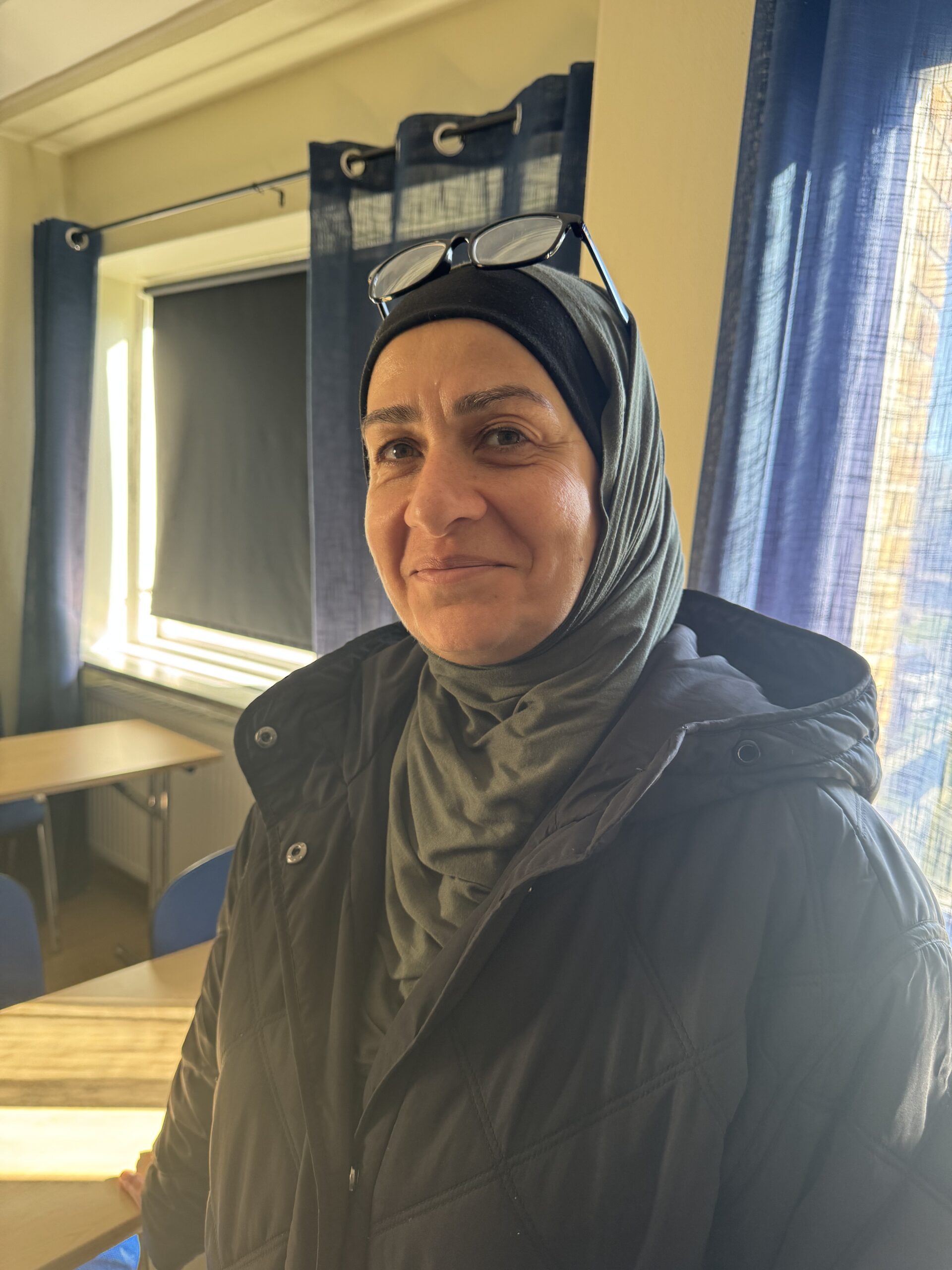 En kvinna med hijab och glasögon på huvudet ler i ett solbelyst rum med blå gardiner.