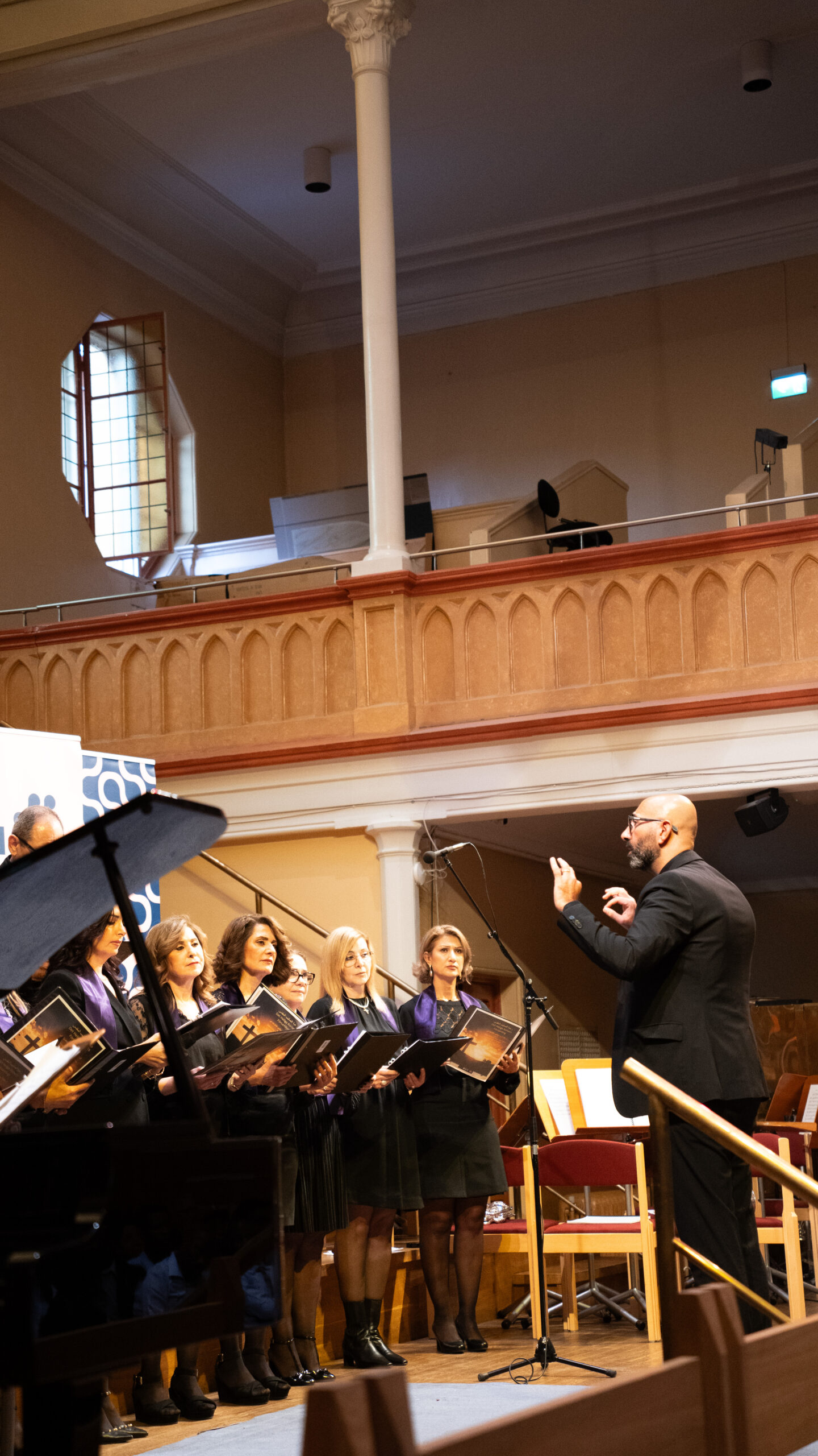 En kvinnokör sjunger inomhus medan en dirigent leder dem med uppsträckta händer.