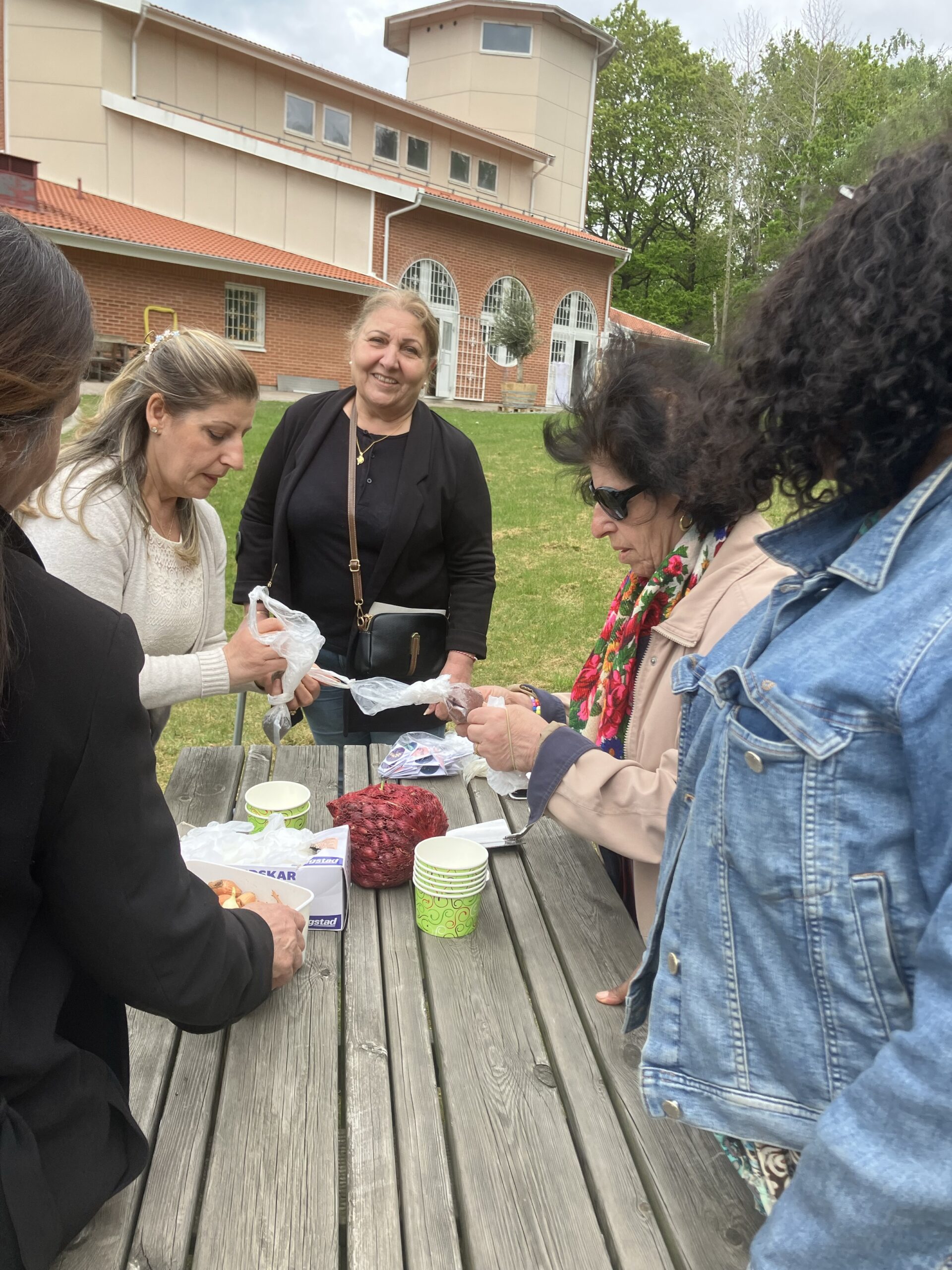 Fem kvinnor samlas runt ett picknickbord utomhus, förbereder mat och ler nära en tegelbyggnad.