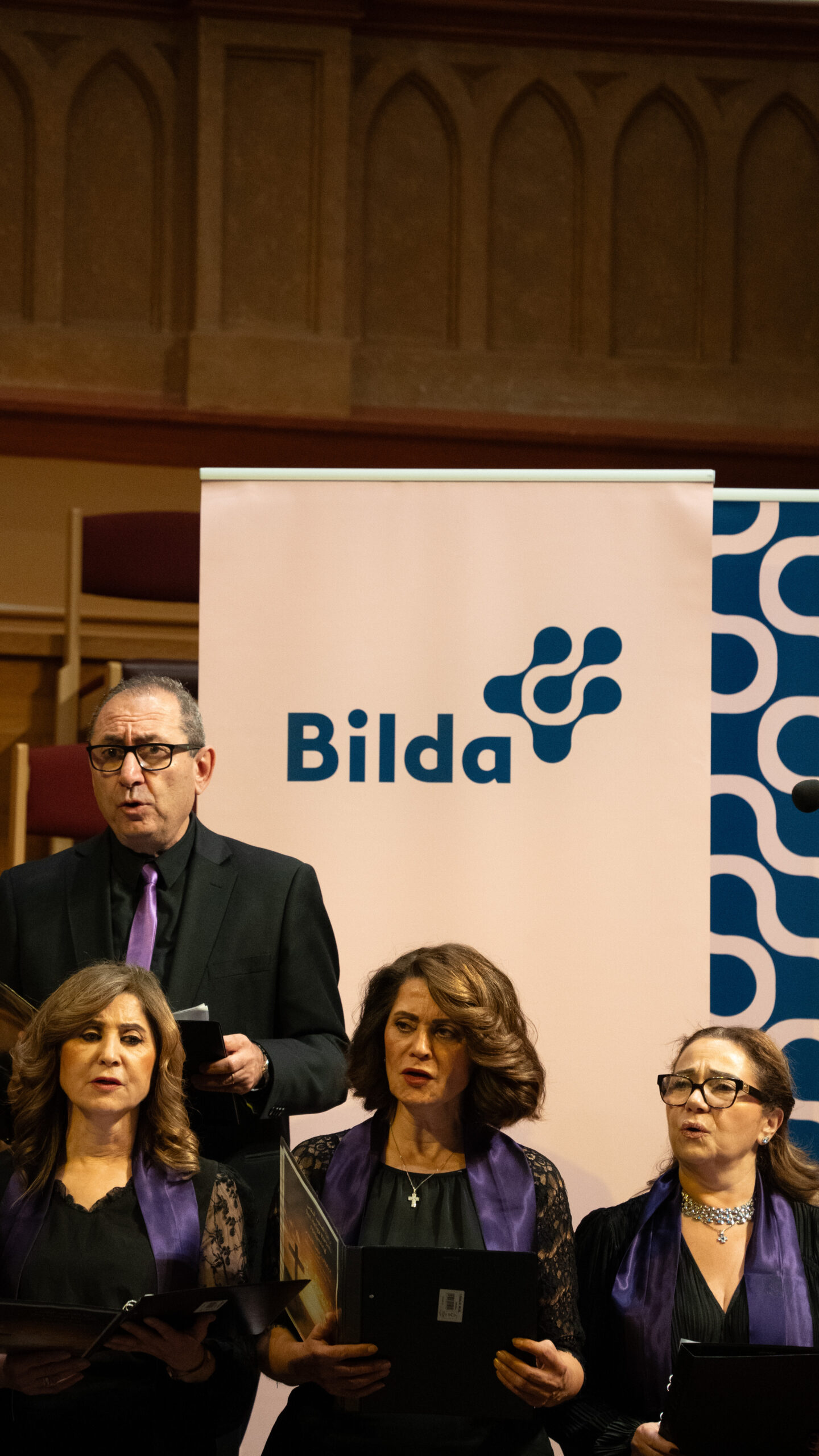 Fyra svartklädda körsångare med lila halsdukar uppträder framför en skylt med texten "Bilda" inomhus.