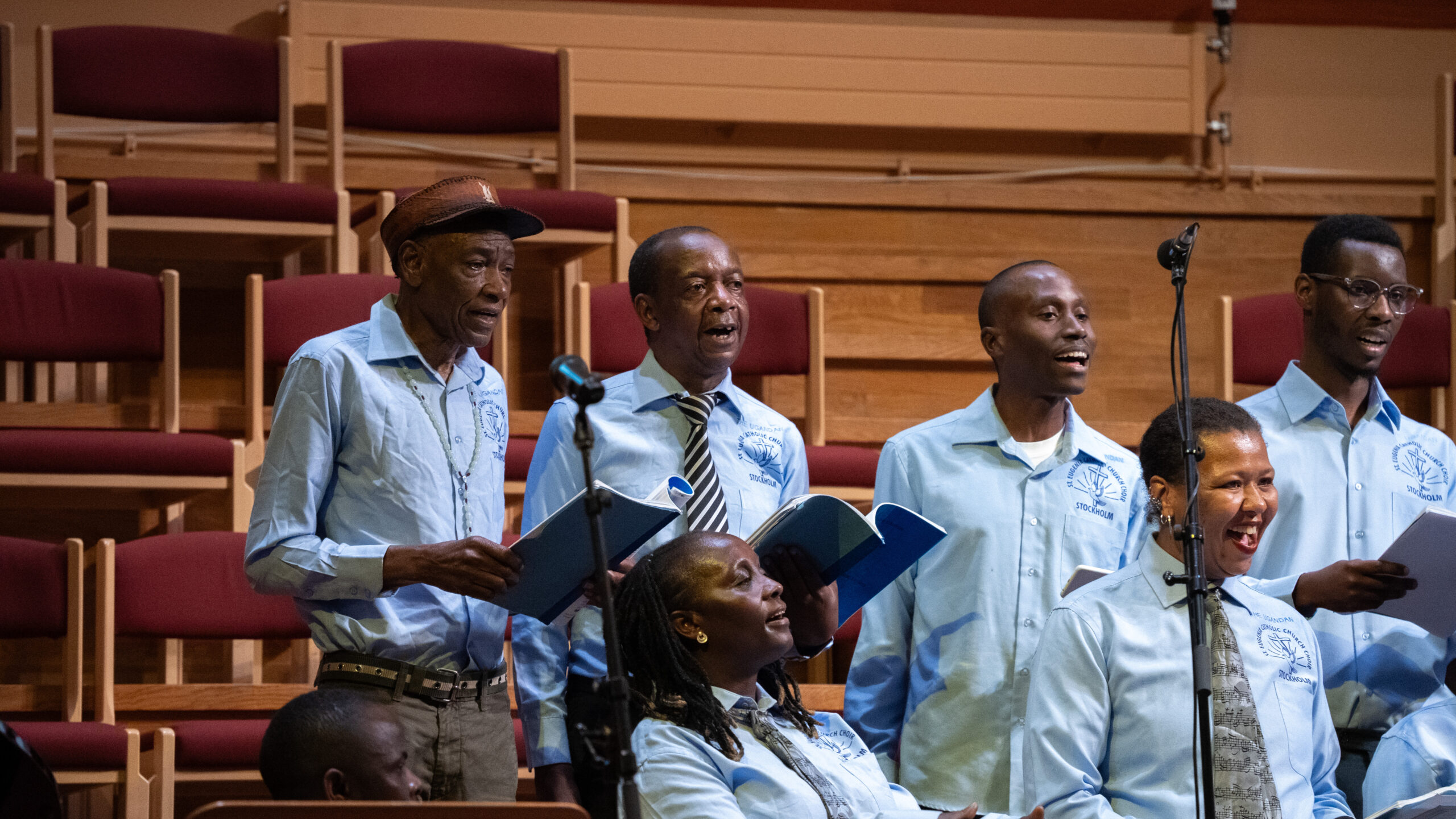 En kör i ljusblå skjortor sjunger tillsammans på scenen, med noter i händerna och leenden på läpparna.