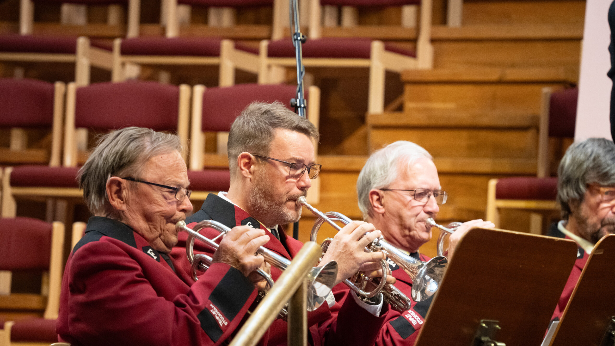 Fyra män i rödbruna kavajer spelar trumpet i en sittande blåsorkester med tomma stolar bakom sig.