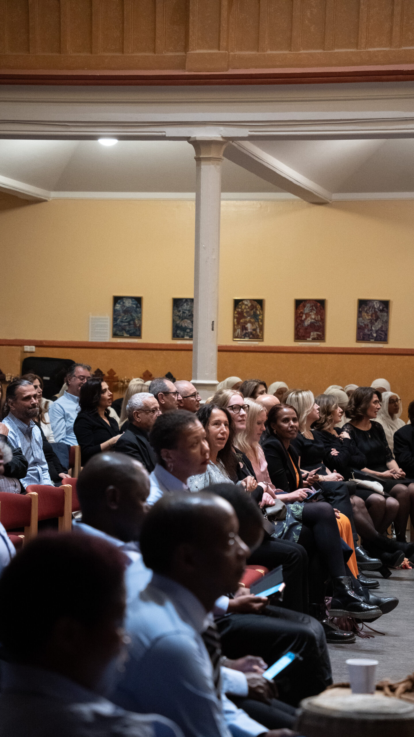 Människor sitter i rader och följer uppmärksamt en händelse i en kyrka eller en sal med inramade bilder på väggen.