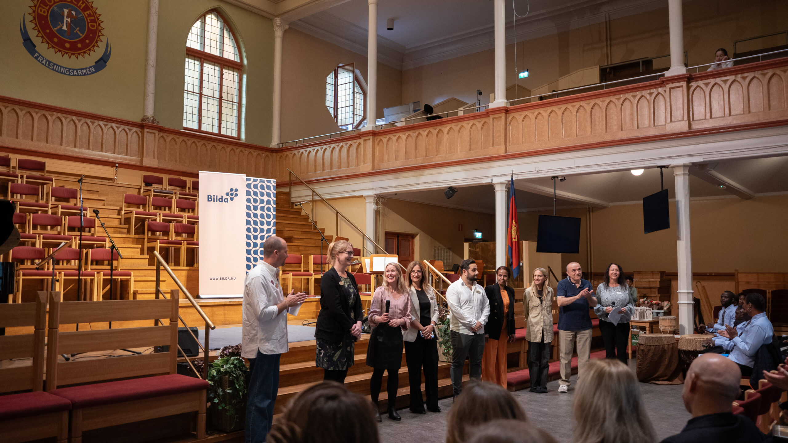 En grupp människor står på scenen i en stor sal, vända mot en publik, under ett evenemang inomhus.