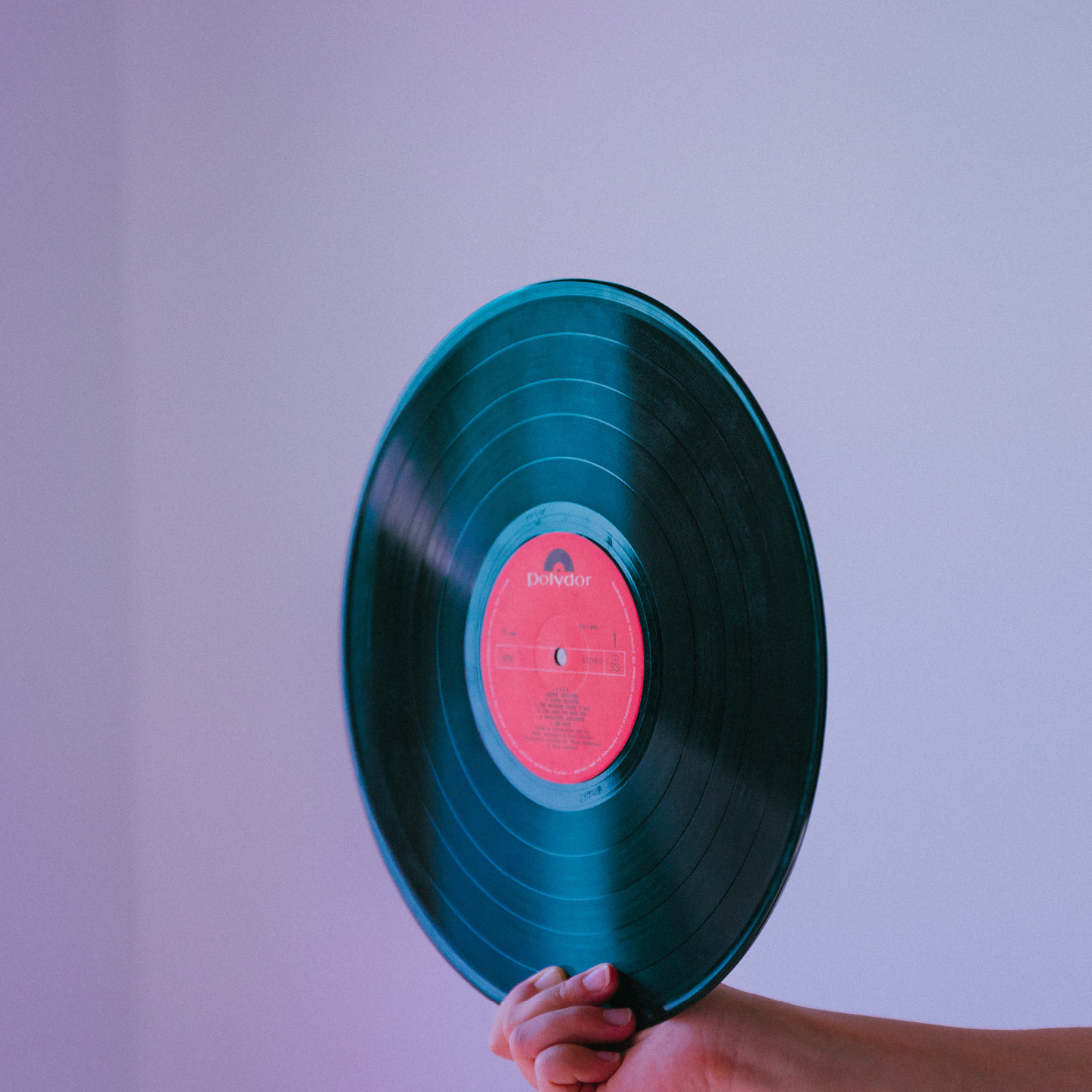 En hand håller upp en vinylskiva med röd etikett mot en enfärgad lila bakgrund.