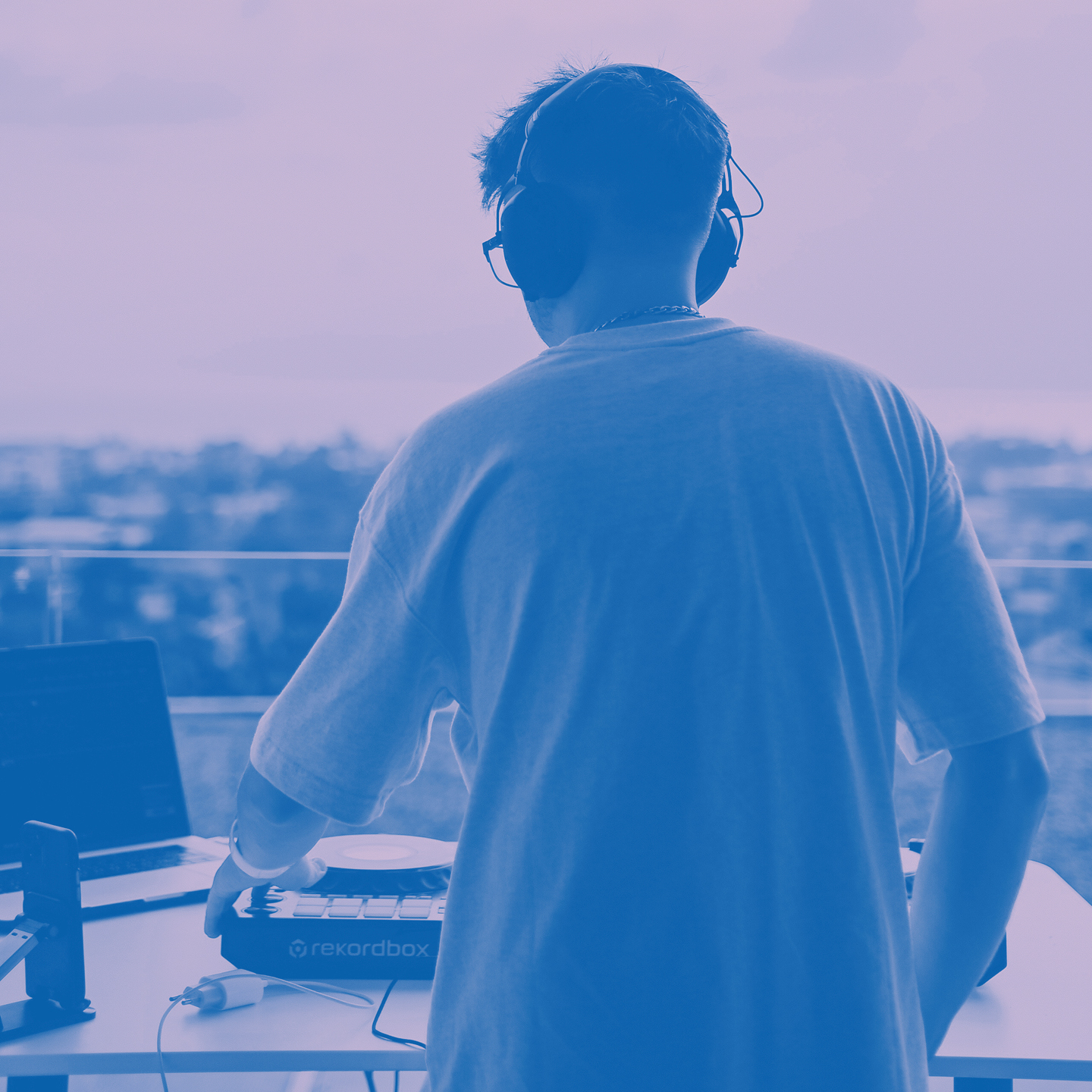 Person med hörlurar mixar musik på en DJ-kontroller bredvid en bärbar dator, vänd mot en utomhusvy.