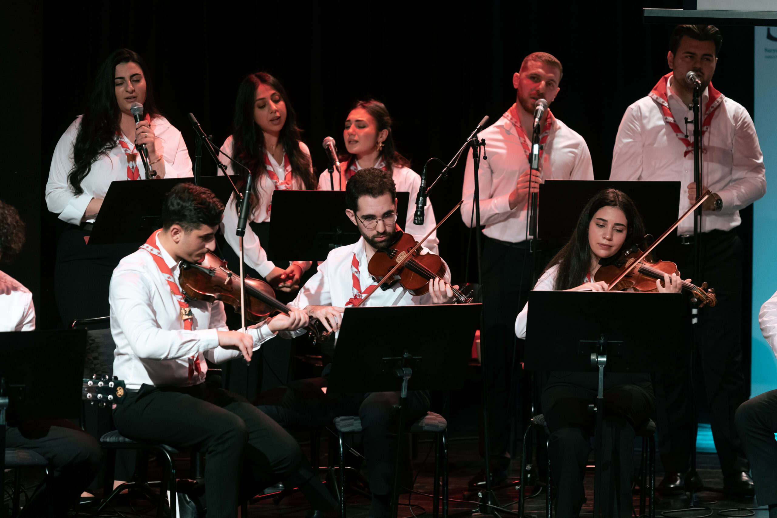 En grupp musiker och sångare uppträder på scenen, klädda i vita skjortor med röda halsdukar.
