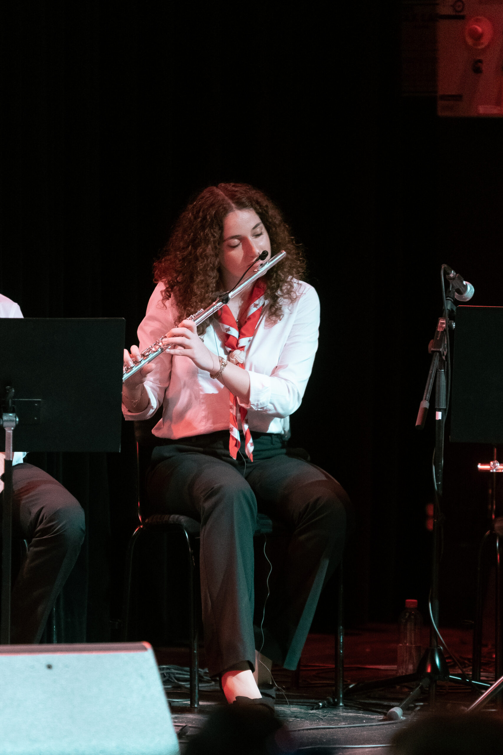 En kvinna med lockigt hår spelar flöjt på scenen, sittande och klädd i vit skjorta med röd halsduk.