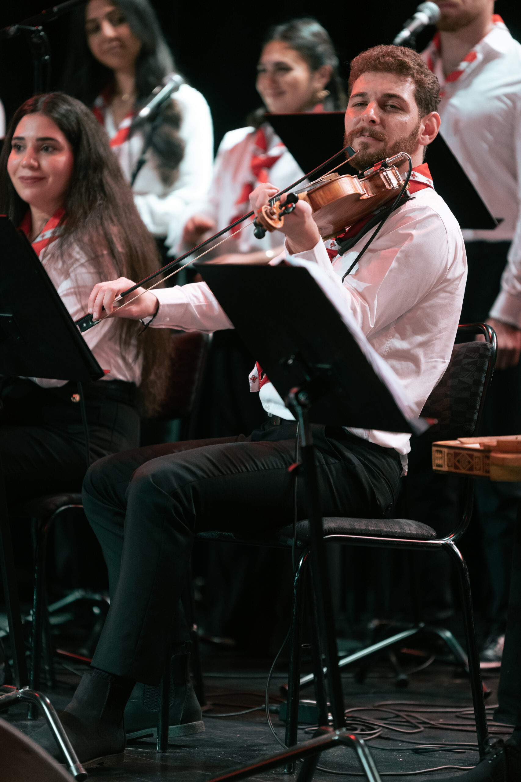 En man i vit skjorta spelar violin på scenen med en sittande kvinna och andra musiker i bakgrunden.