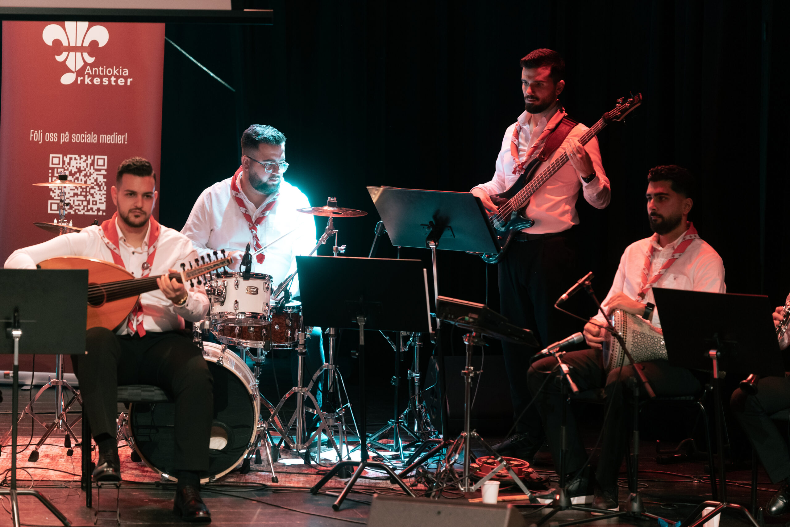 Fyra manliga musiker i vita skjortor spelar instrument på en mörk scen med notställ framför sig.