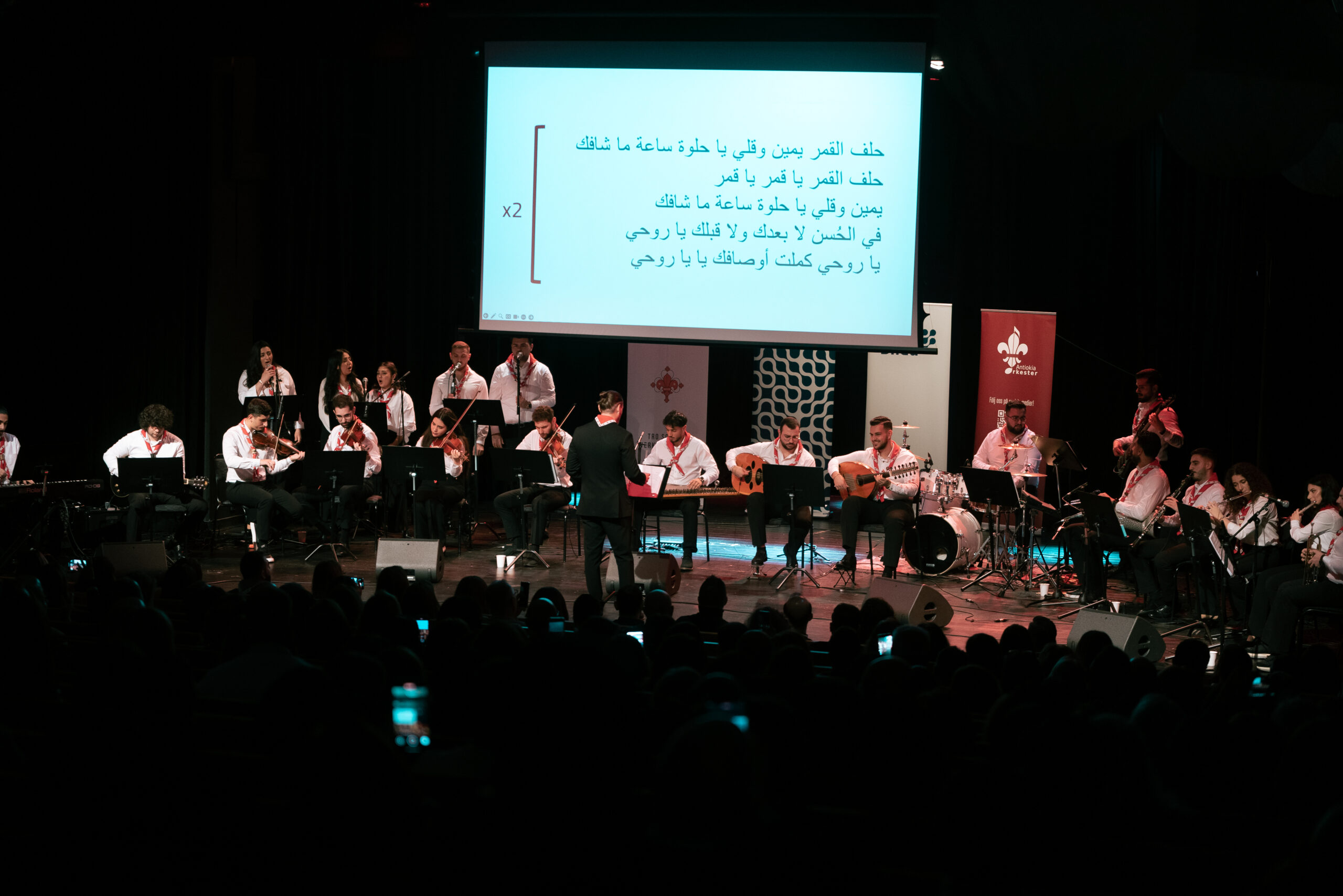 En stor musikensemble uppträder på scenen med en skärm som visar arabisk text bakom dem.