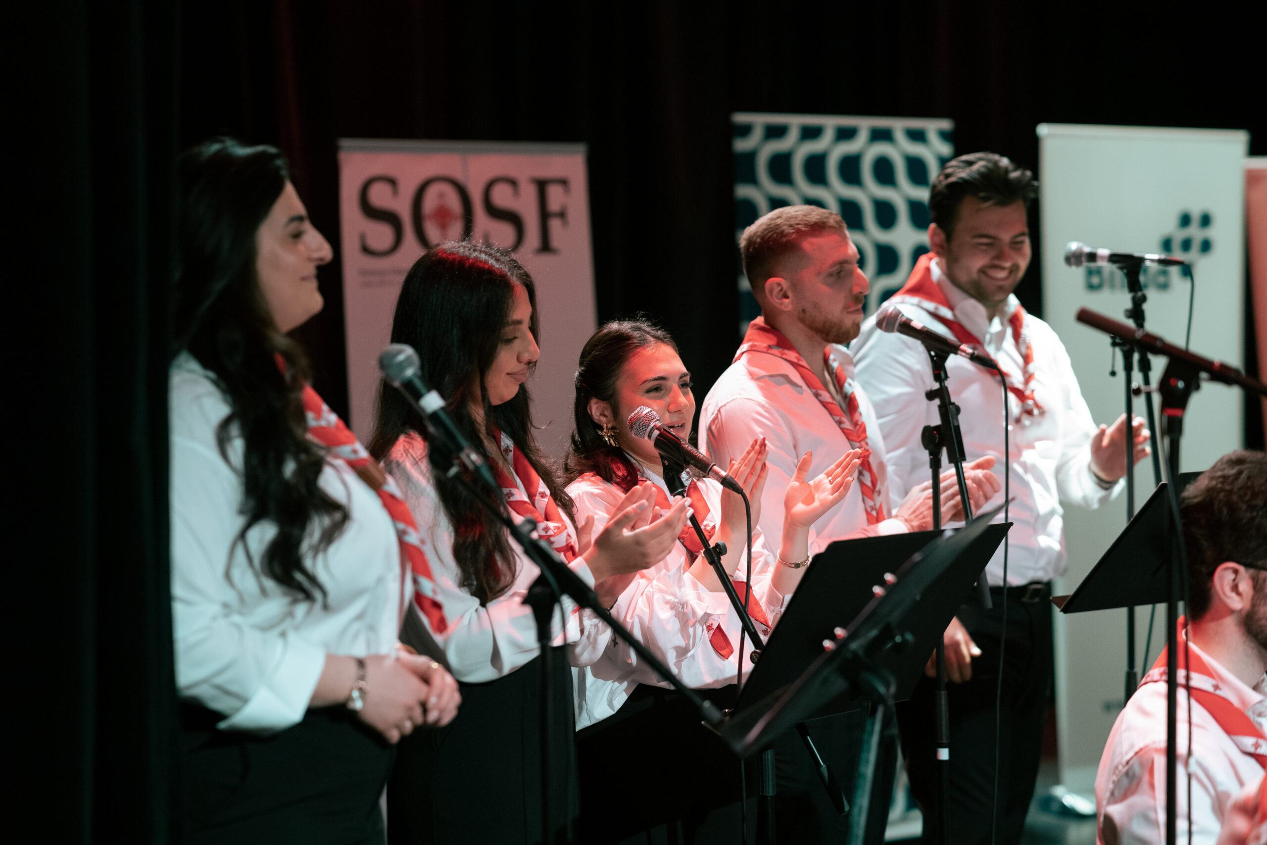Fem personer i matchande dräkter sjunger och klappar på scenen med mikrofoner och banderoller i bakgrunden.