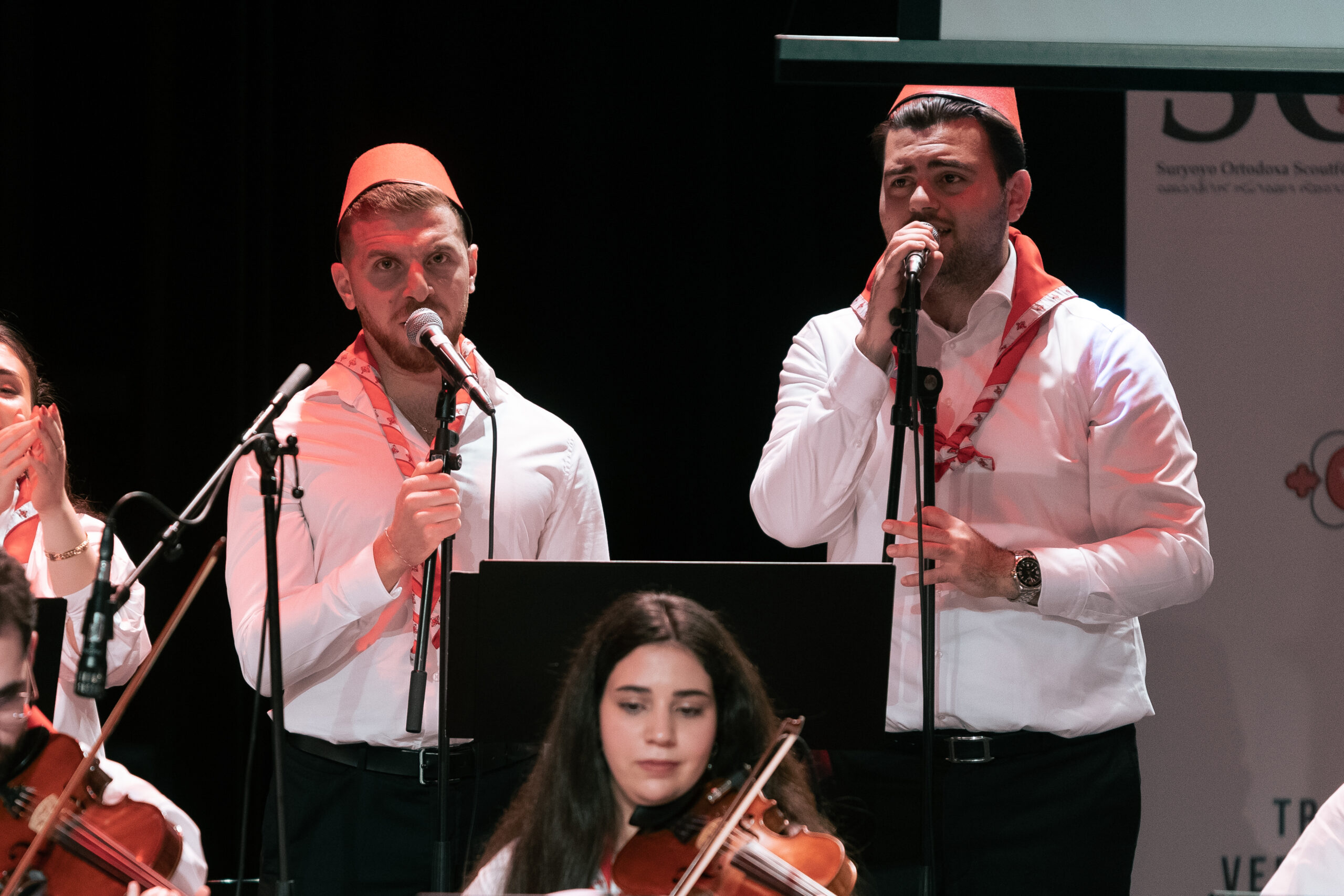 Två män i vita skjortor och röda hattar sjunger i mikrofoner på scenen; en kvinna spelar violin framför dem.