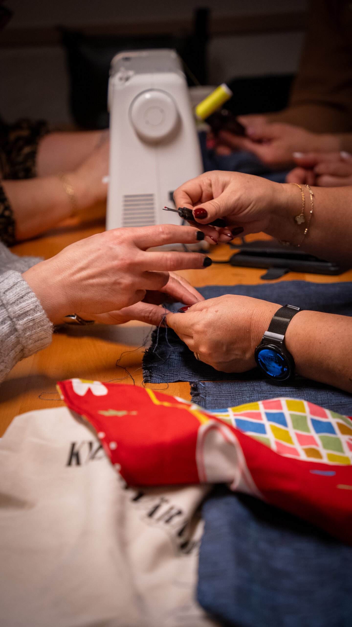 Tre personer syr ihop tyg för hand, med en symaskin och färgglada tyger på bordet.